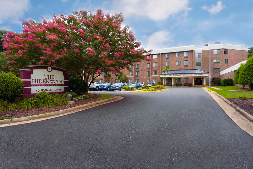 Exterior view of The Hidenwood Retirement Community building with a driveway leading to the entrance. There is a large tree with pink flowers and a sign that reads 'The Hidenwood Retirement Community, 50 Wellesley Drive' near the driveway. Several cars are parked near the building under a partly cloudy sky.