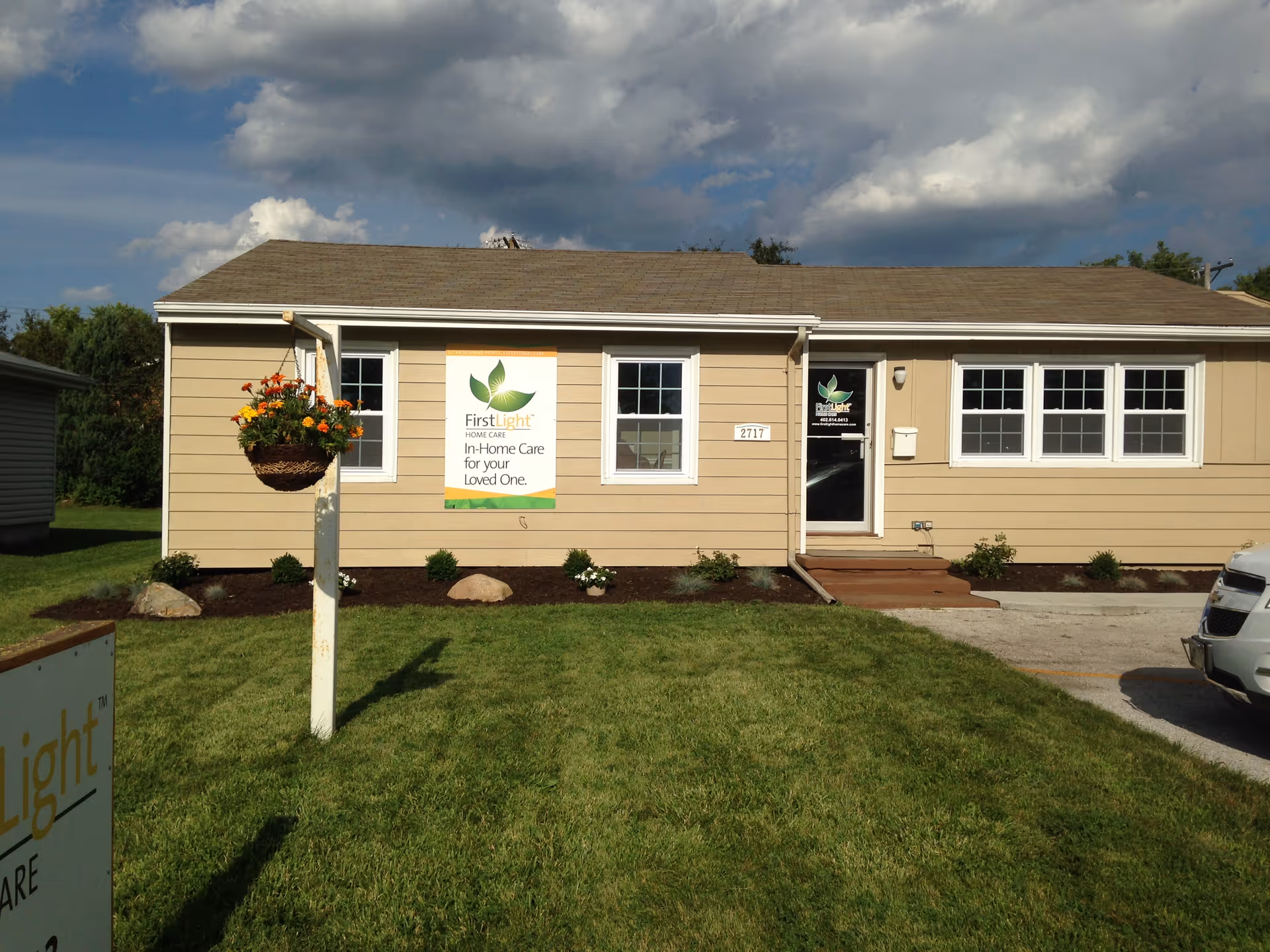 Single-story beige facility with FirstLight Home Care sign, front lawn, and hanging flower basket.