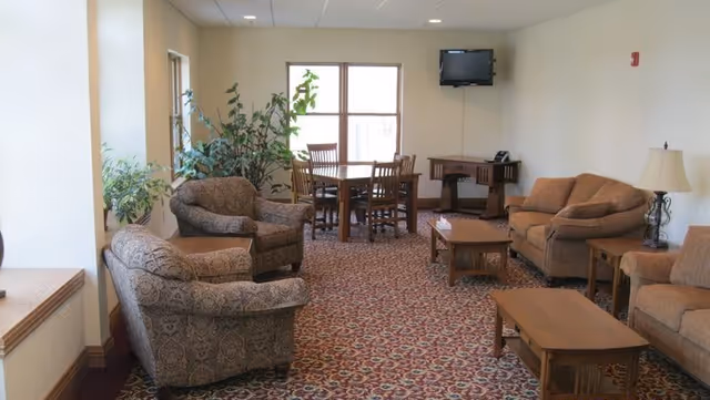 Carpeted senior living lounge with sofas, chairs, tables, plants, and a wall-mounted TV.