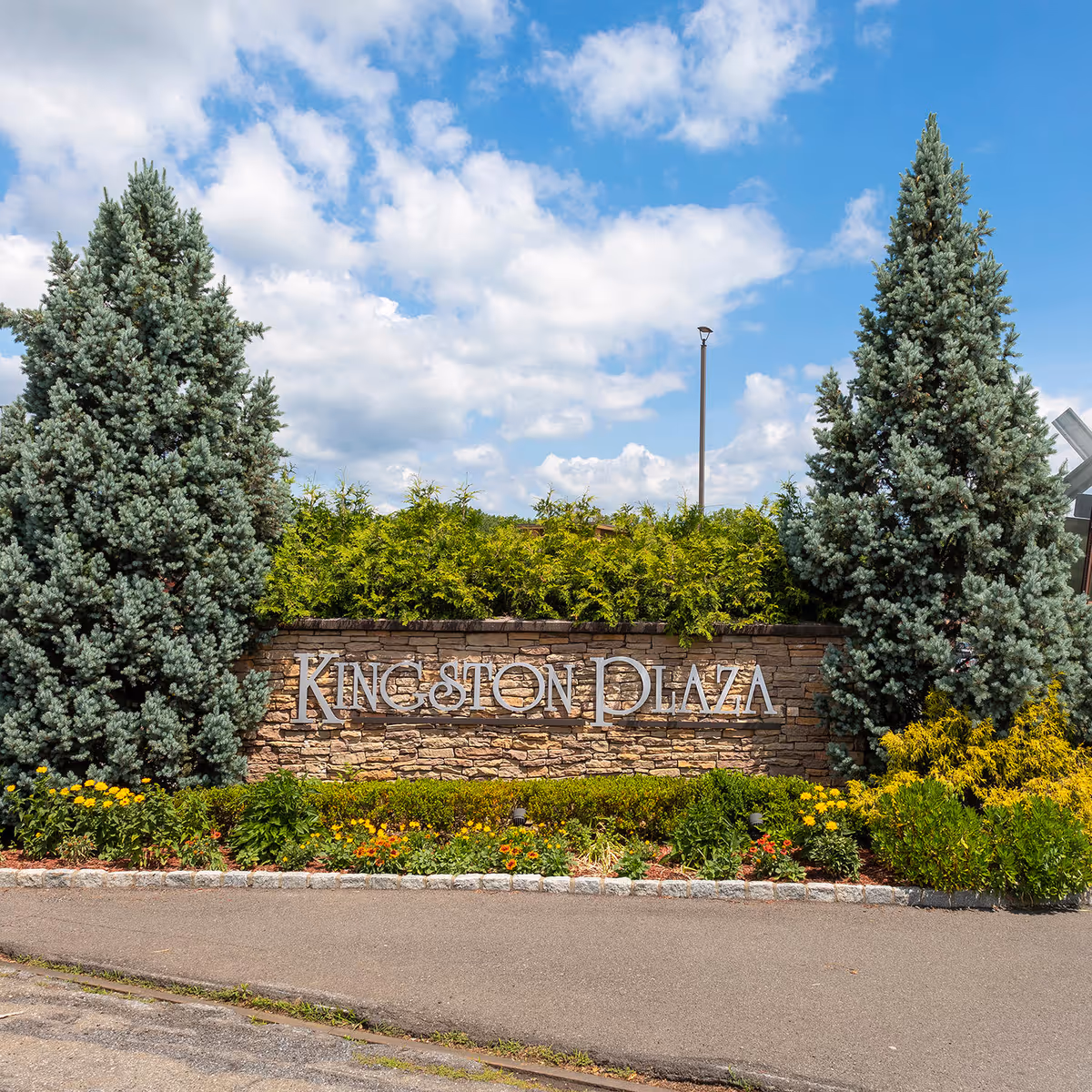 Stone Kingston Plaza sign with greenery and tall evergreens under a blue sky.