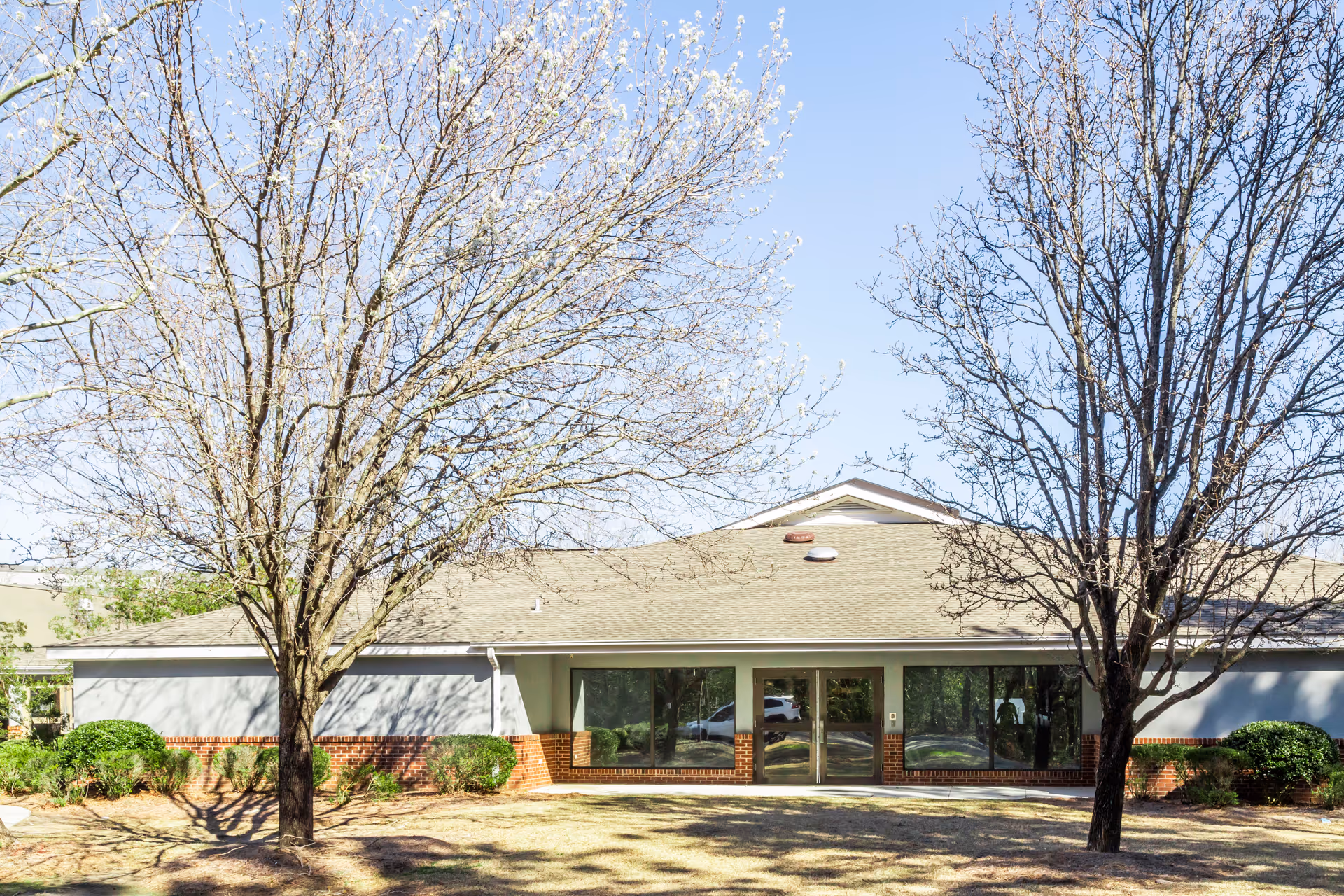 Exterior view of a single-story building with a light gray roof and brick accents at the base. The building has large windows and a double glass door entrance. In front of the building are two leafless trees and some green shrubs under a clear blue sky.