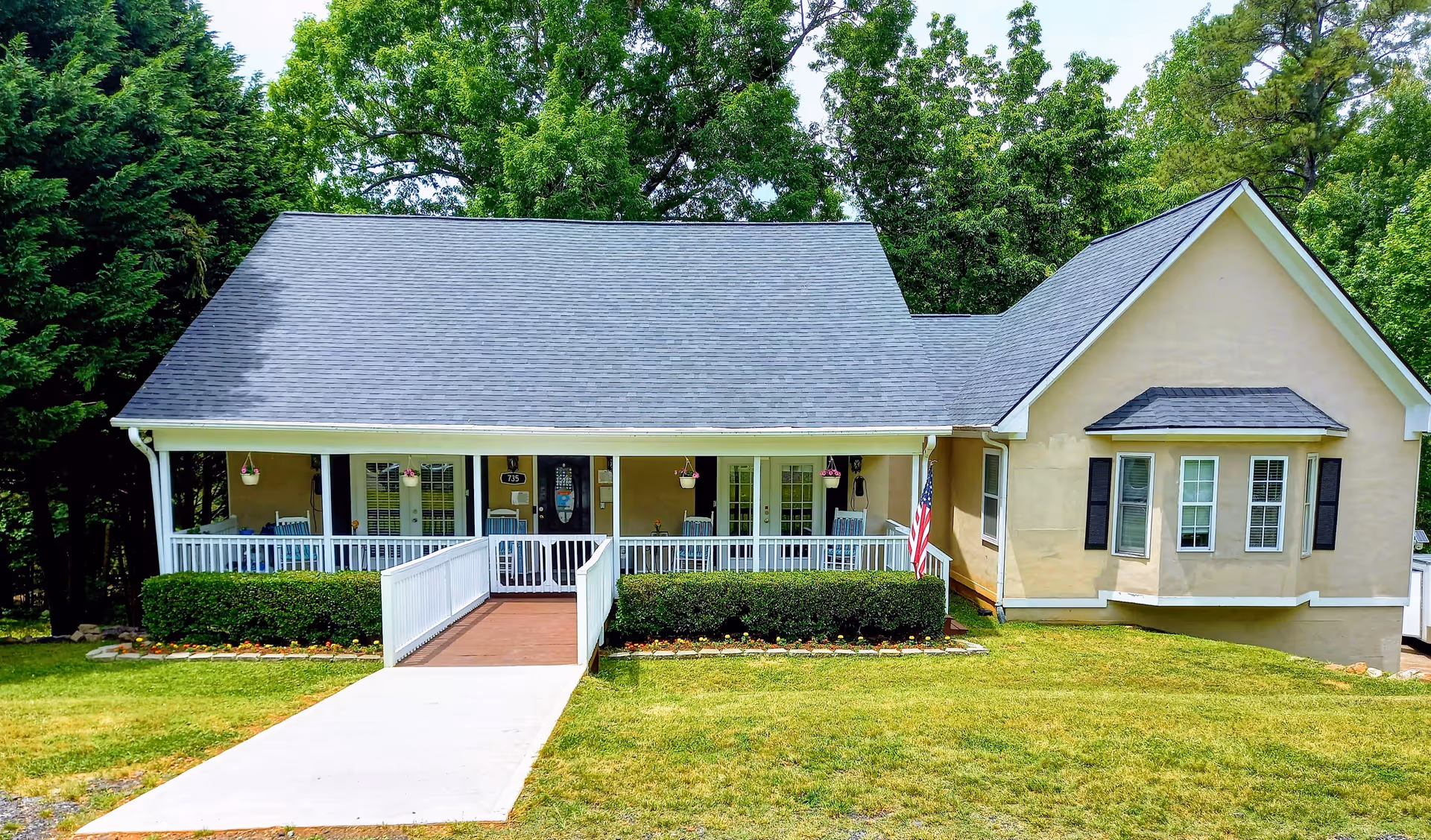 Light-colored single-story building with a broad covered front porch, wheelchair ramp, and American flag on a grassy lawn.