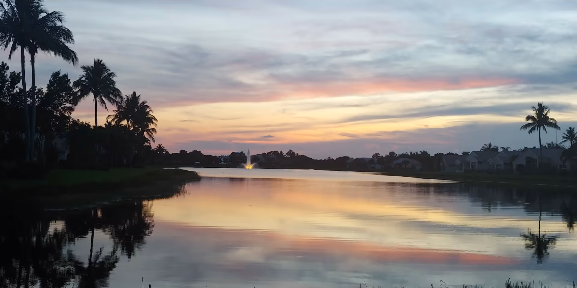 Sunset over a calm lake with palm trees and distant houses.