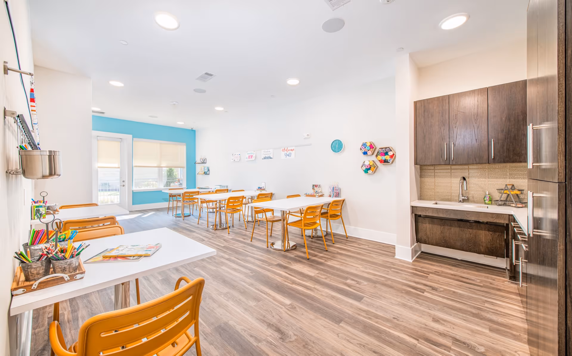 Bright and spacious activity room with white walls and wood flooring, featuring several white tables with orange chairs arranged for group activities. A small kitchenette with dark wood cabinets and a sink is on the right side. Art supplies and colorful decorations are visible on the tables and walls, including hexagonal shelves with yarn and a blue clock. Large windows with blinds allow natural light to fill the room.