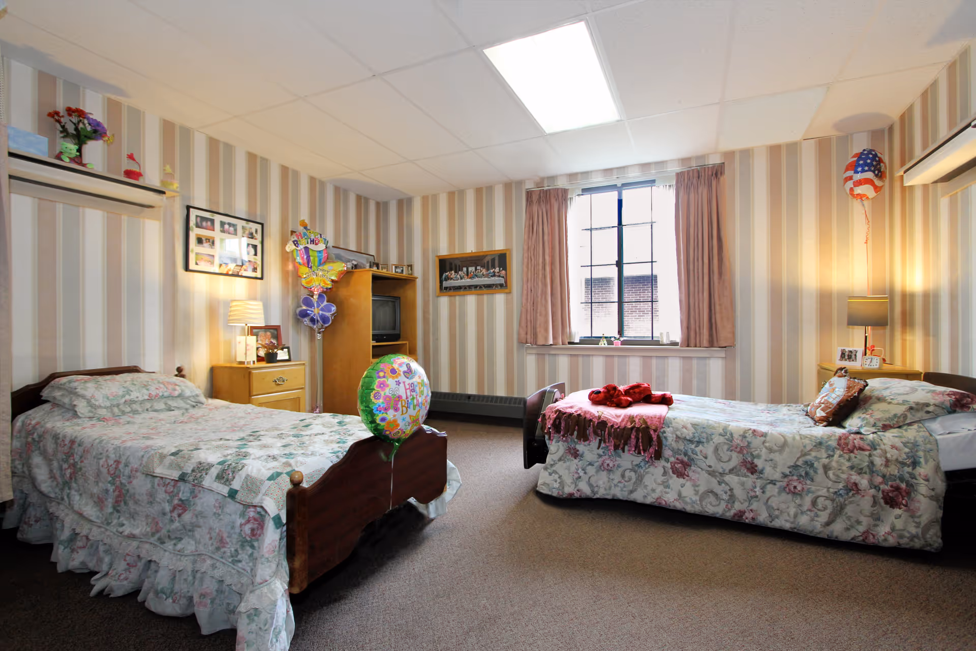 Two twin beds with floral bedspreads in a decorated shared bedroom with striped wallpaper, a TV cabinet, window and balloons.