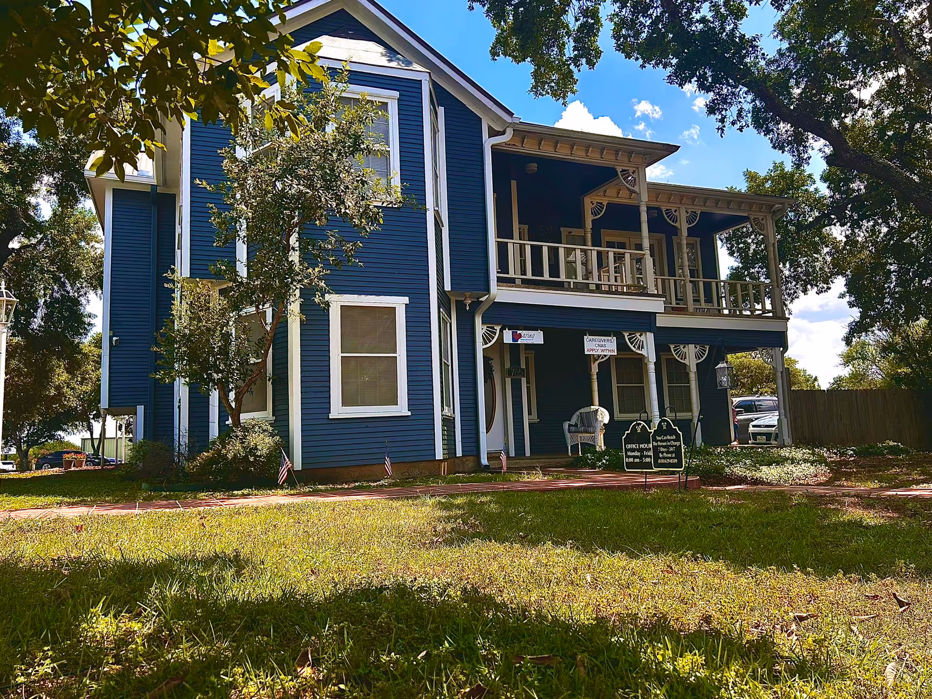 Blue two-story senior care building with a wraparound porch and lawn in front.