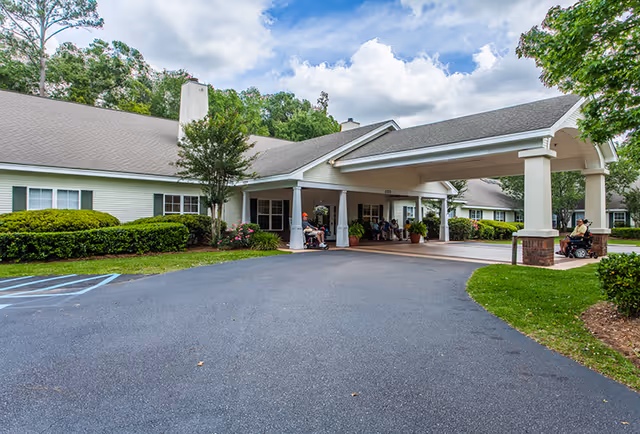 Exterior view of Oaks at Charleston senior living facility showing a covered entrance with columns, a paved driveway, green bushes, trees, and a few people sitting outside near the entrance.