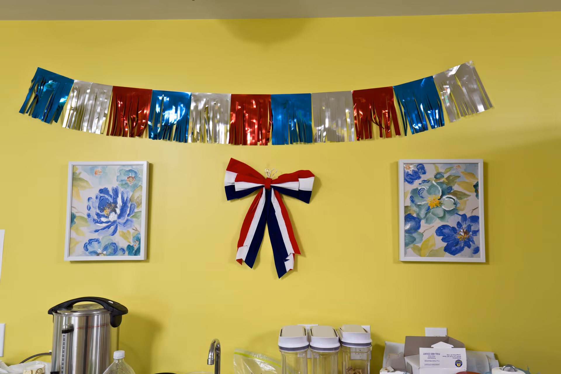 A yellow wall decorated with a red, white, and blue bow in the center and a garland of metallic red, blue, and silver fringe above it. Two framed floral paintings with blue flowers are hung on either side of the bow. Below the decorations, there is a countertop with a stainless steel appliance, clear containers with white lids, and some papers.