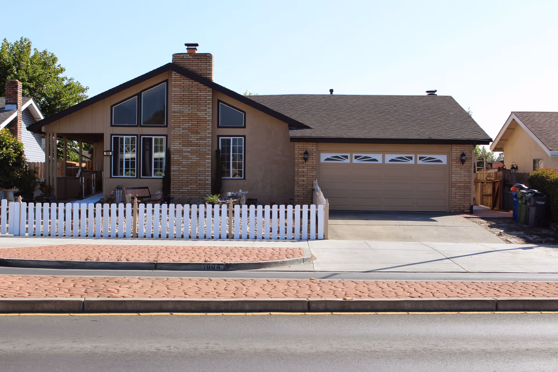 Front exterior view of a single-story house with a brick chimney, large windows, a two-car garage, and a white picket fence in front. The house is situated along a street with a sidewalk and a patterned brick pedestrian crossing area.