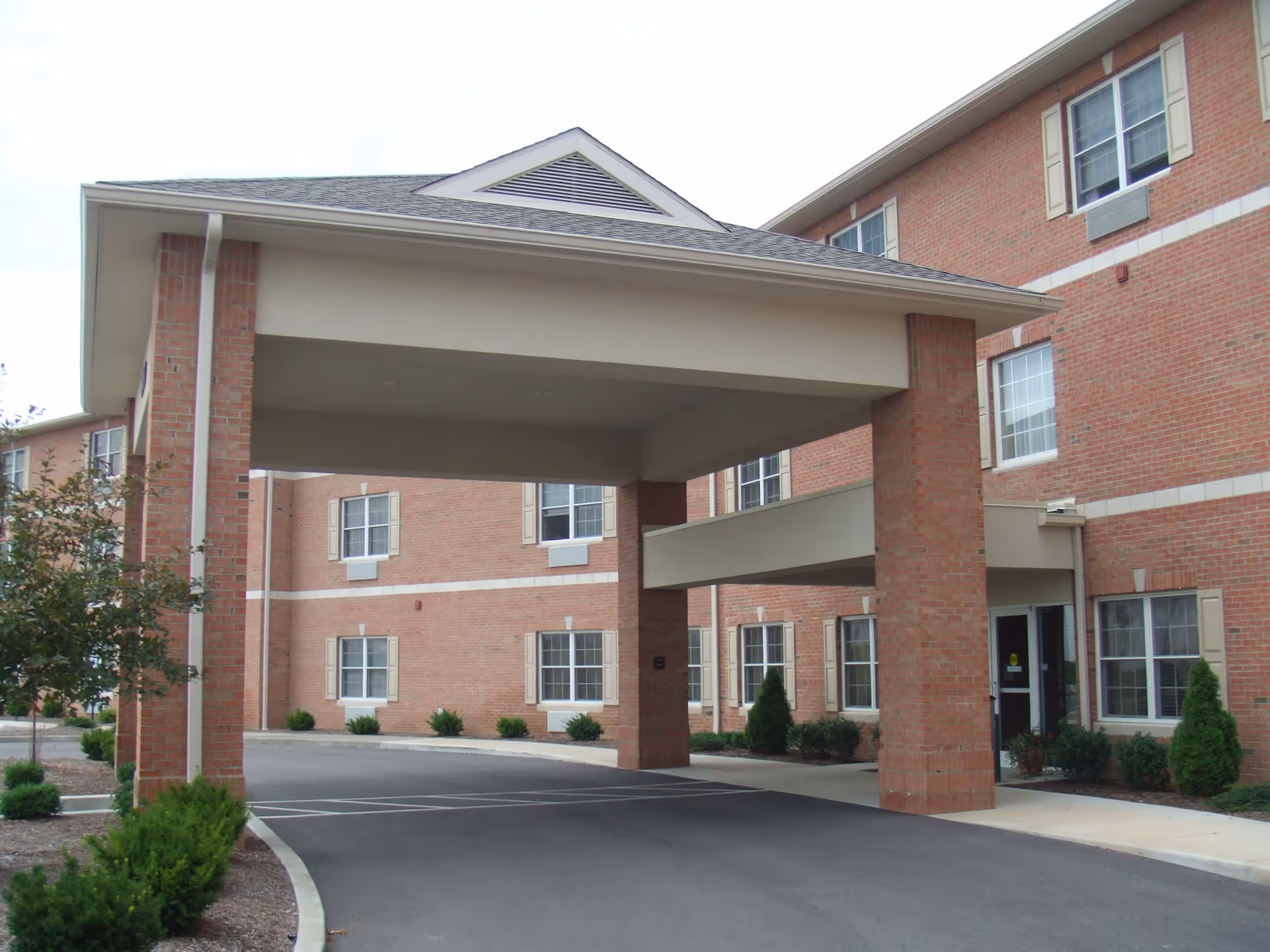 Covered entrance area of a brick building with multiple windows and beige trim, surrounded by small shrubs and a paved driveway.