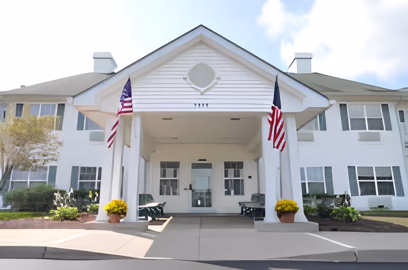 Front entrance of a white two-story senior living building with a covered portico flanked by American flags and potted flowers.