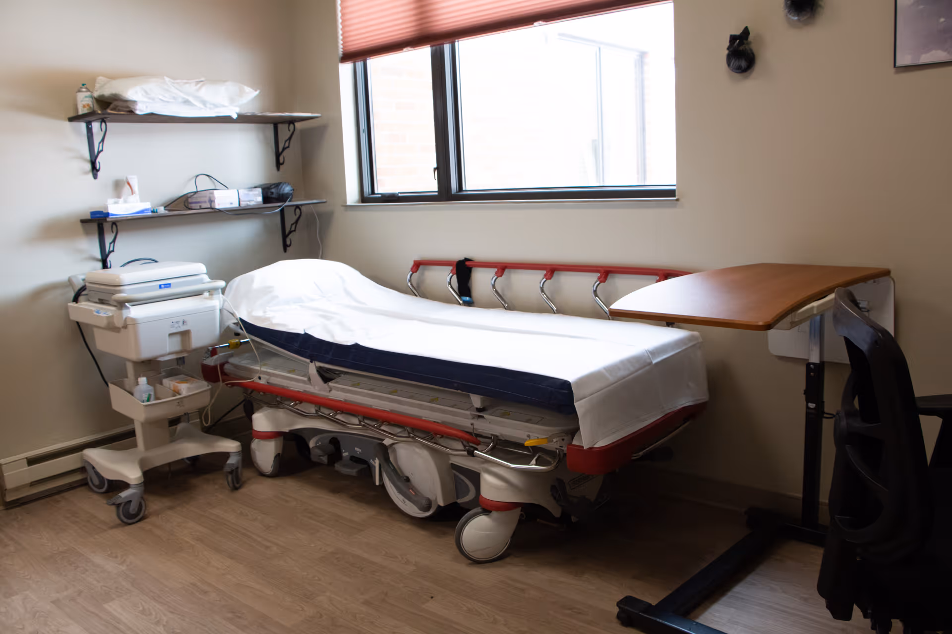 A hospital room with a medical bed covered with white sheets, a rolling medical cart with supplies, two wall-mounted shelves holding various items, a window with a red blind, a wooden overbed table, and a black chair.
