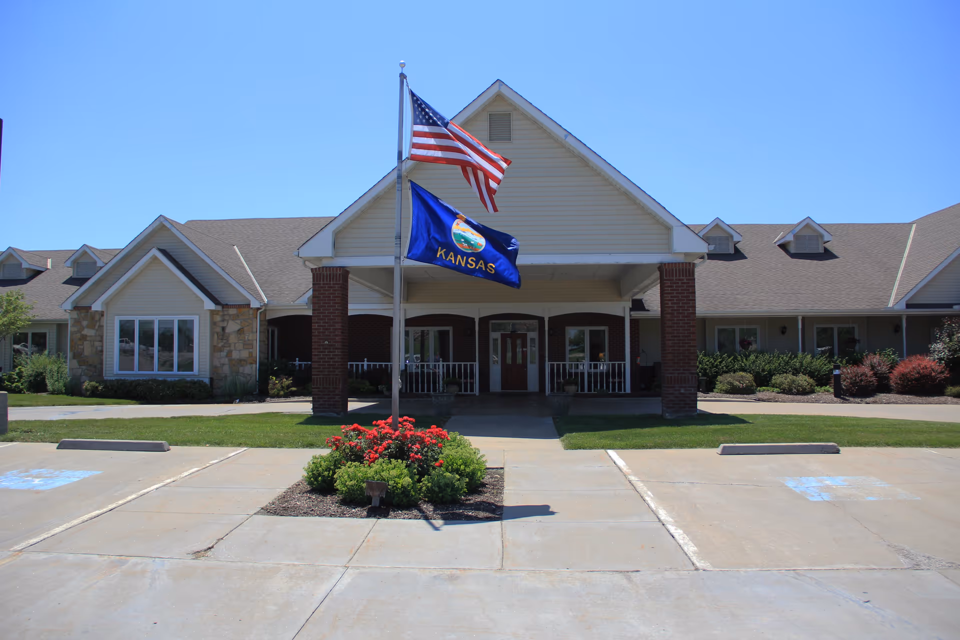 Front exterior view of Vintage Park at Baldwin City building with a covered entrance supported by brick columns, an American flag and Kansas state flag flying on flagpoles, and a small landscaped area with red flowers in front of the entrance. The sky is clear and blue.