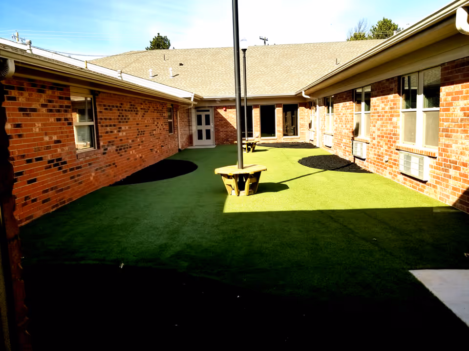 Sunlit brick courtyard with artificial turf, picnic-style benches around a light pole, and surrounding building windows and doors.