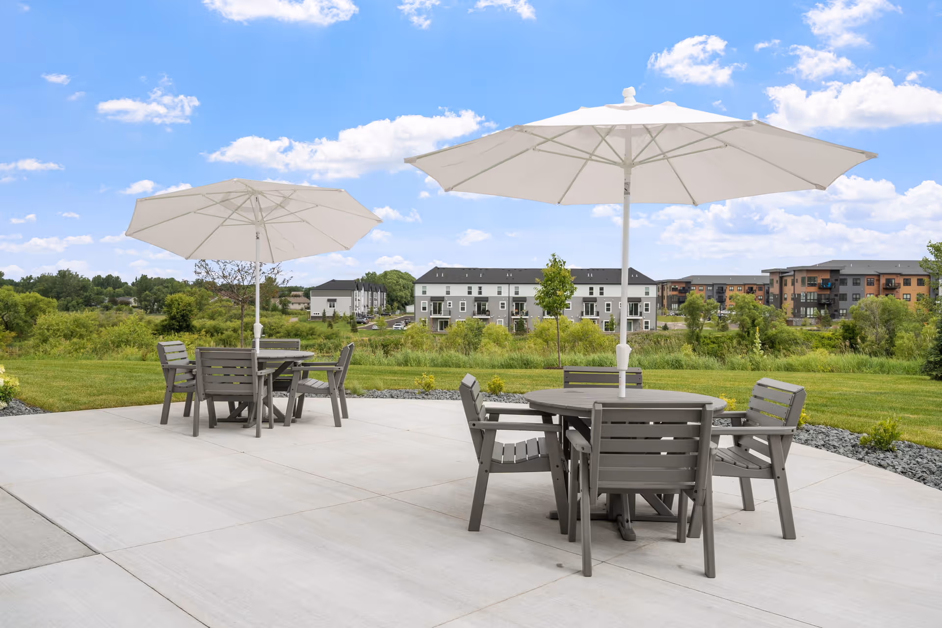 Outdoor patio with tables, chairs, and umbrellas overlooking a grassy area and residential buildings.