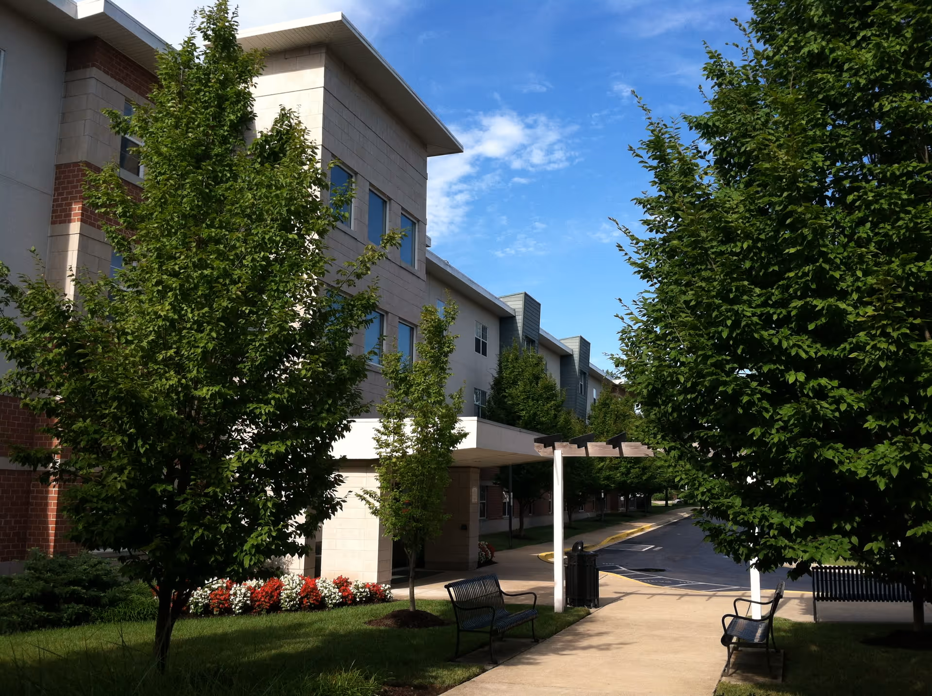 Exterior view of Oaks at Liberty facility showing a multi-story building with large windows, surrounded by green trees and landscaped flower beds. There is a paved walkway with benches and a pergola structure, under a blue sky with some clouds.