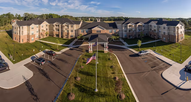 Aerial front view of a large three-story senior living building with a circular driveway, flagpoles, parked cars, and landscaped lawns.
