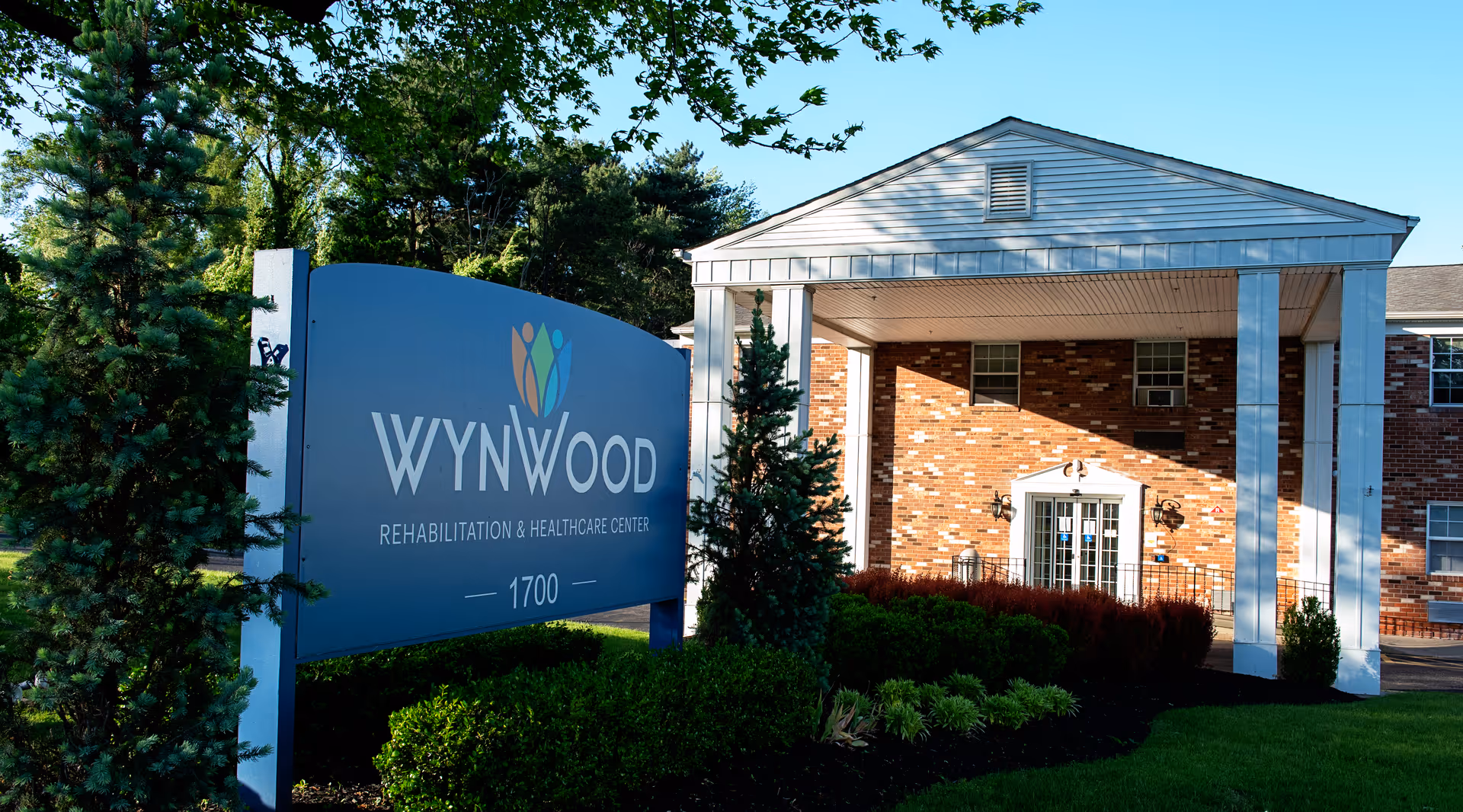 Exterior view of Wynwood Rehabilitation and Healthcare Center building with a large blue sign displaying the facility's name and address, surrounded by green trees and shrubs under a clear blue sky.