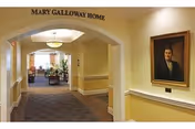Indoor hallway in a senior living facility with arched entry, seating area, and wall portrait.