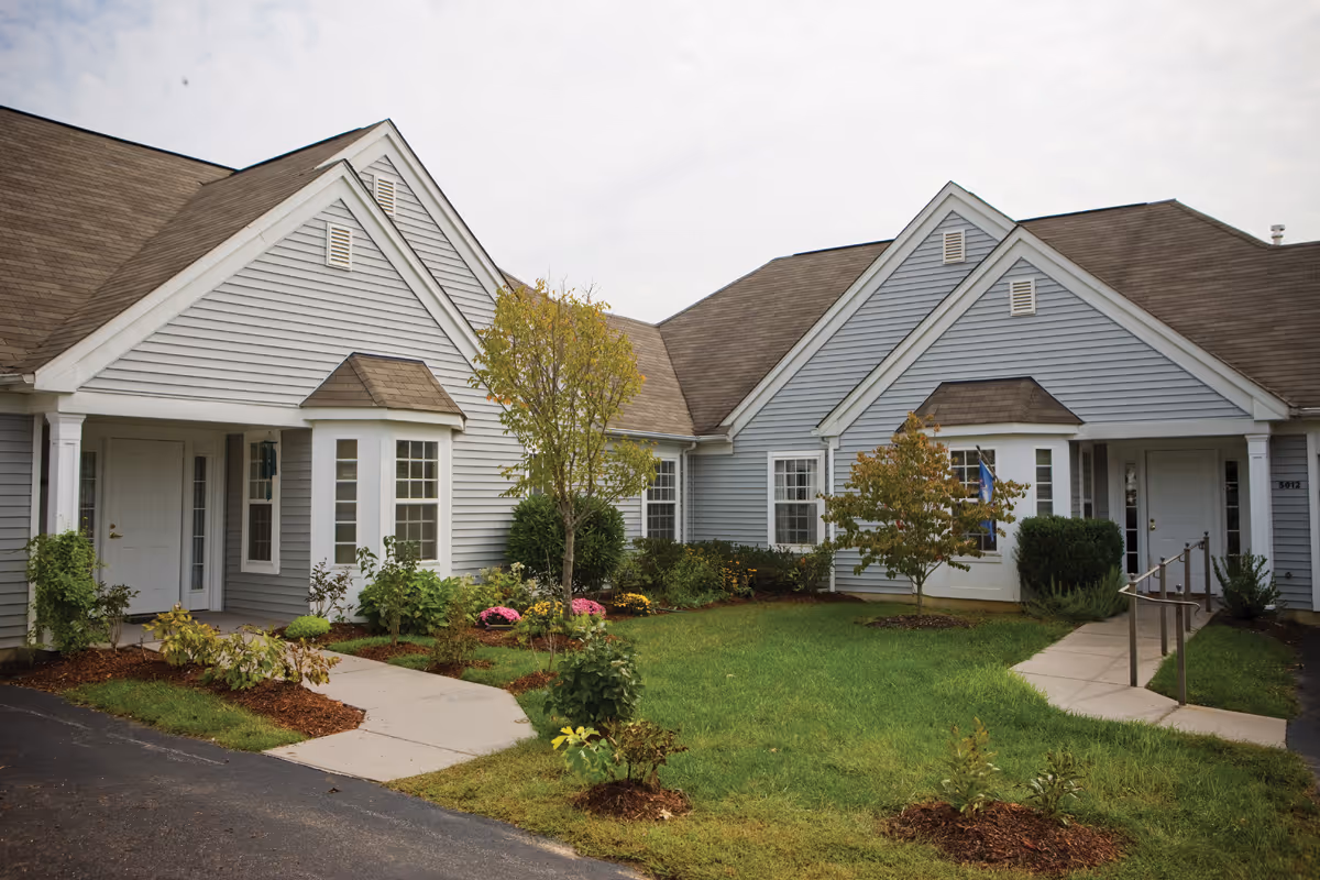 Exterior view of a senior living facility building with gray siding, white trim, and brown shingled roofs. The building has multiple peaked roof sections and several windows. There is a well-maintained lawn with small trees, shrubs, and flower beds in front of the building. A paved walkway leads to the entrances.