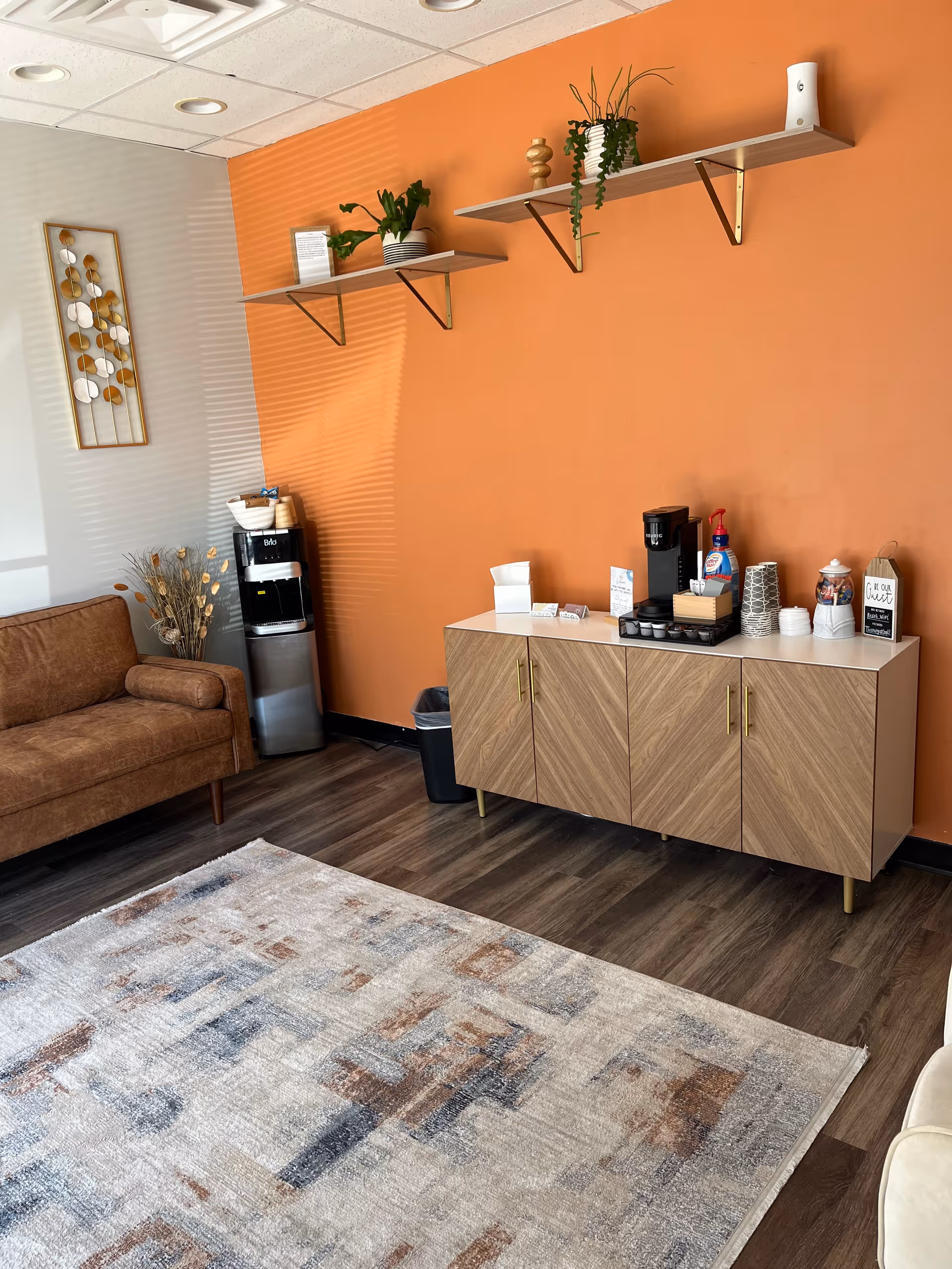 Cozy interior lounge featuring orange wall, shelves with plants, coffee station, and sofa.