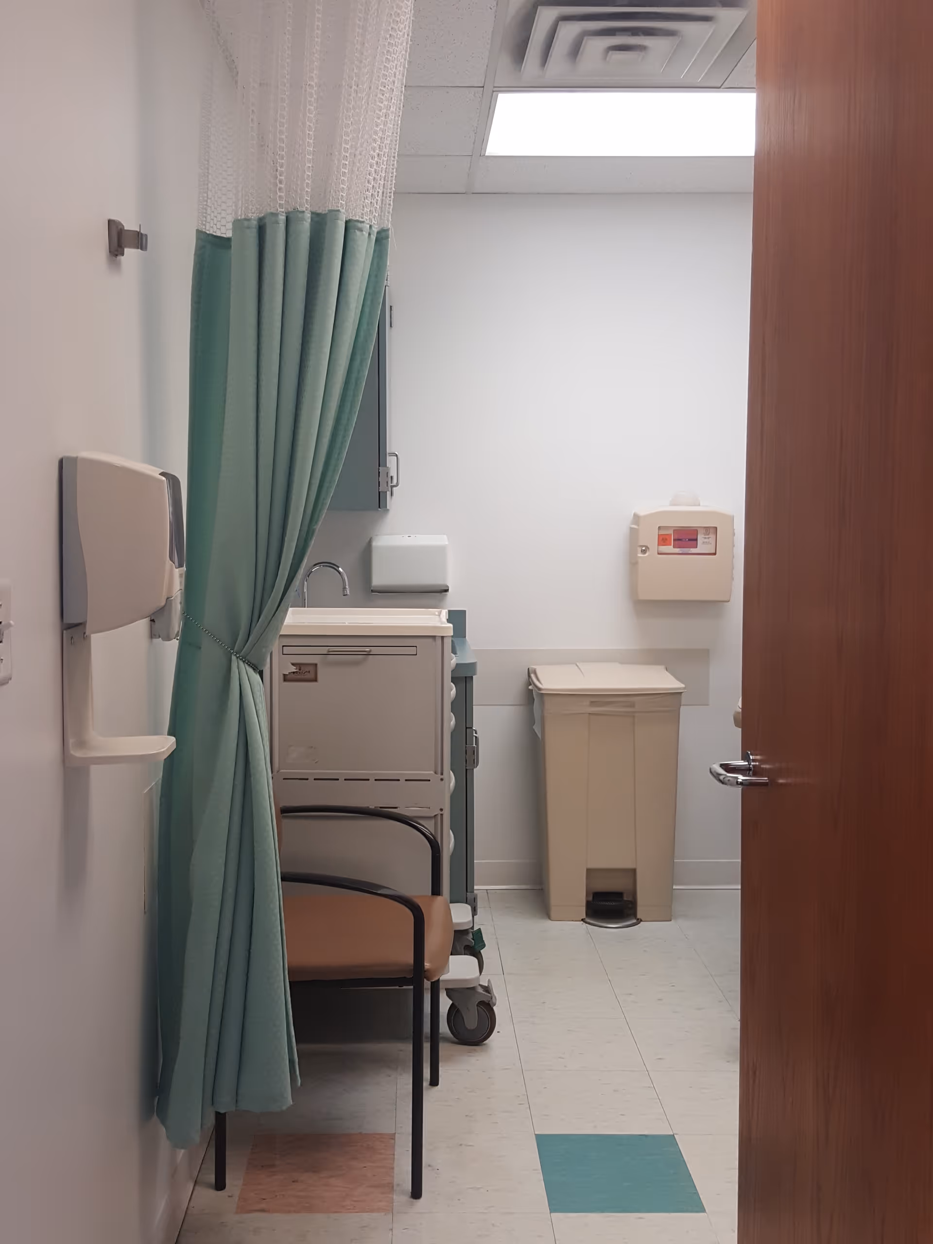 Medical examination room with teal privacy curtain, chair, sink, and trash can.