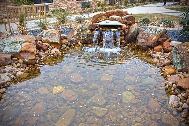 A small outdoor water feature with clear water flowing over rocks into a shallow pond surrounded by stones and landscaping, with a wooden bench and a building visible in the background.