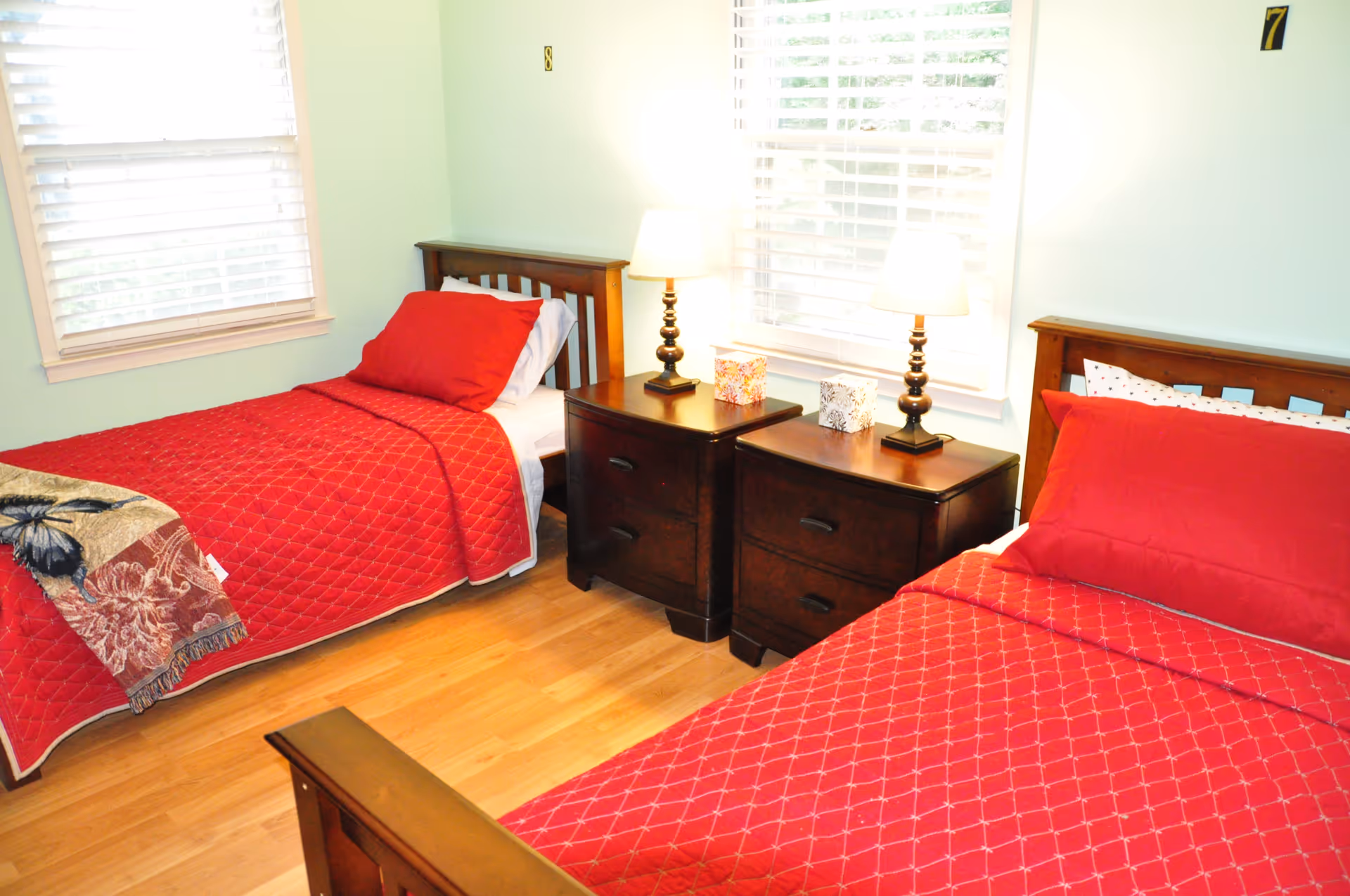 Two single beds with red quilts and matching nightstands in a bright bedroom.