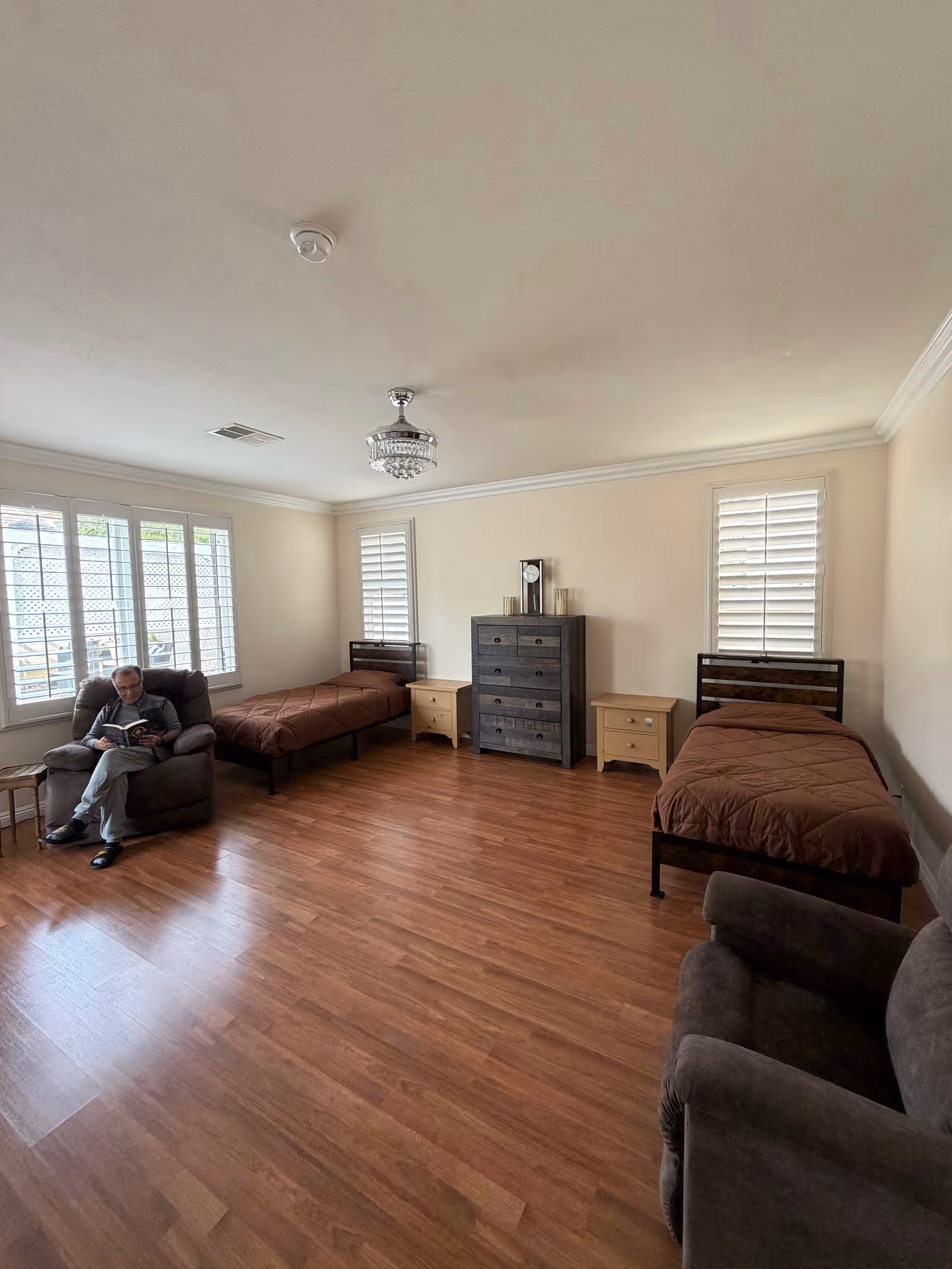 A spacious bedroom in a senior care home with two beds and a man reading.