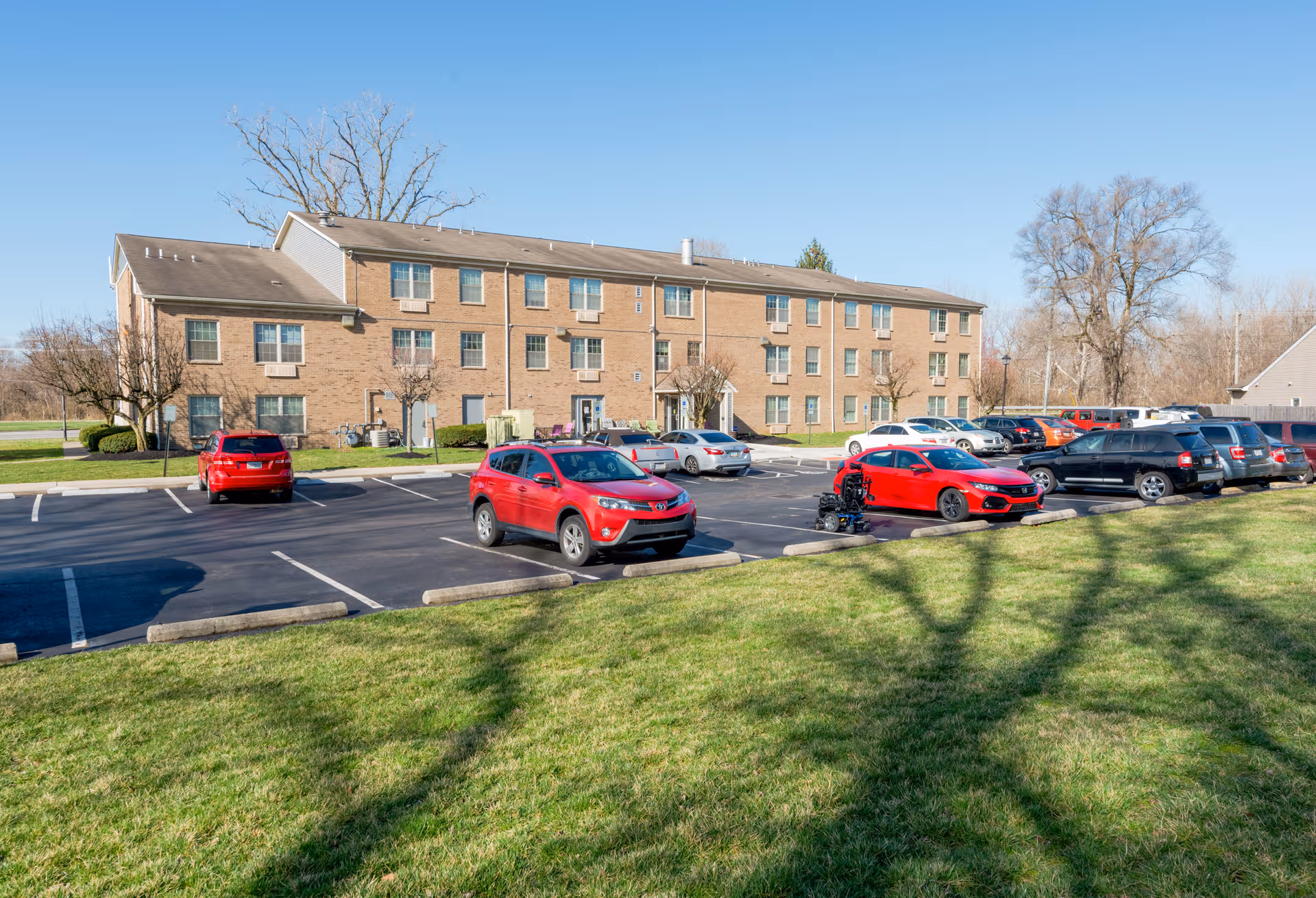 Brick senior living building with a large parking lot and green lawn in the foreground.