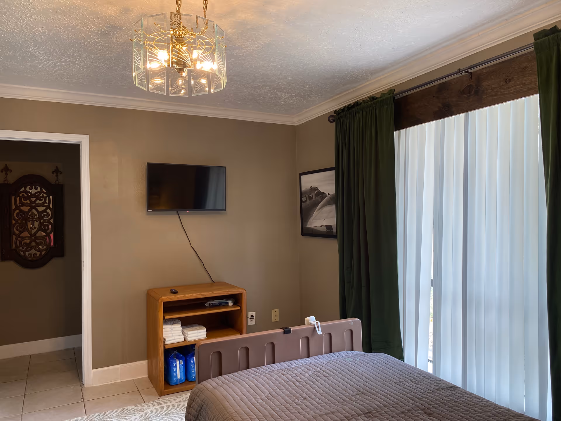 A furnished bedroom with a bed, wall-mounted TV, cabinet, and large window with green curtains.