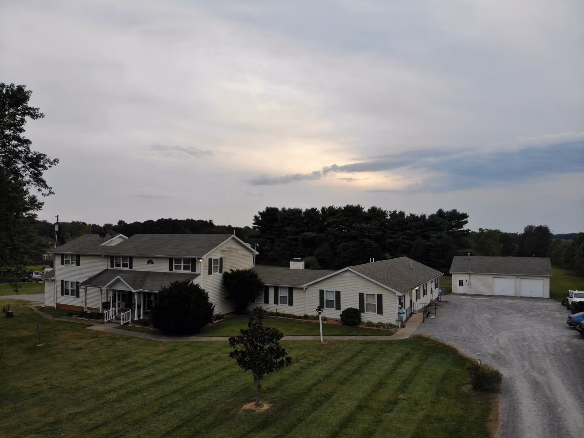 A large two-story residential building with an attached single-story wing, surrounded by a well-maintained lawn and trees. There is a gravel driveway leading to a detached garage with three doors. The sky is cloudy with a hint of sunlight breaking through.