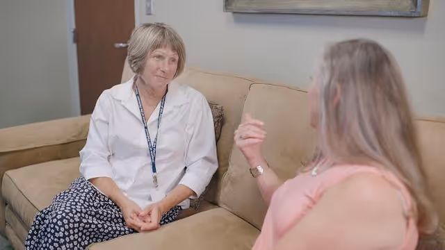 Two women sit on a beige sofa in a living room, engaged in conversation.