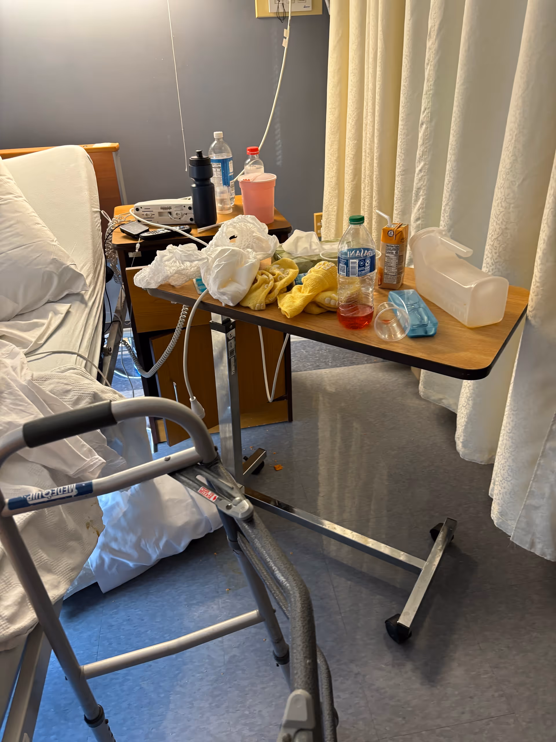 Hospital-style senior care room with bed, walker, cluttered bedside table, and privacy curtain.