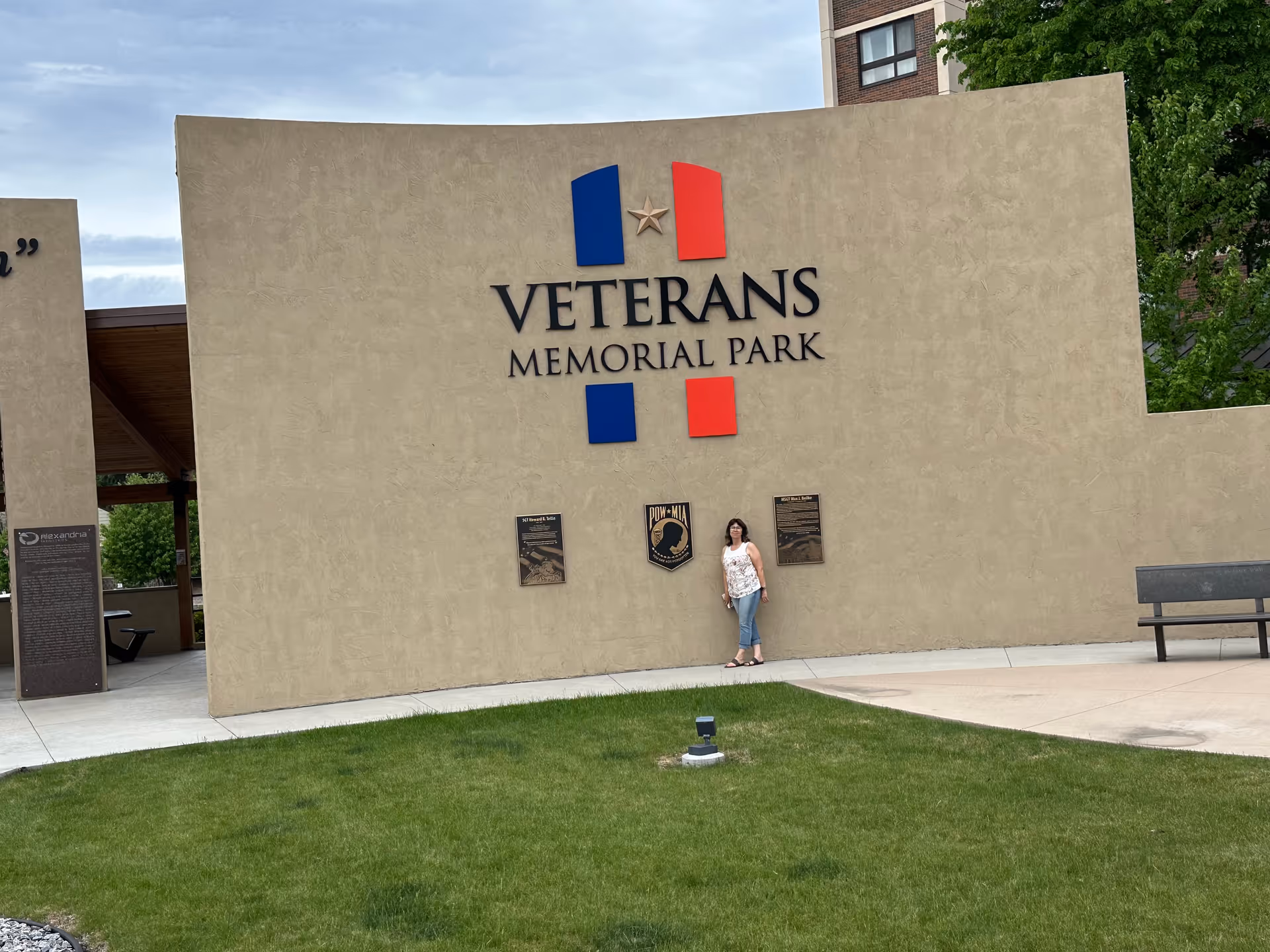 Person standing beside a beige Veterans Memorial Park wall sign on a grassy foreground.