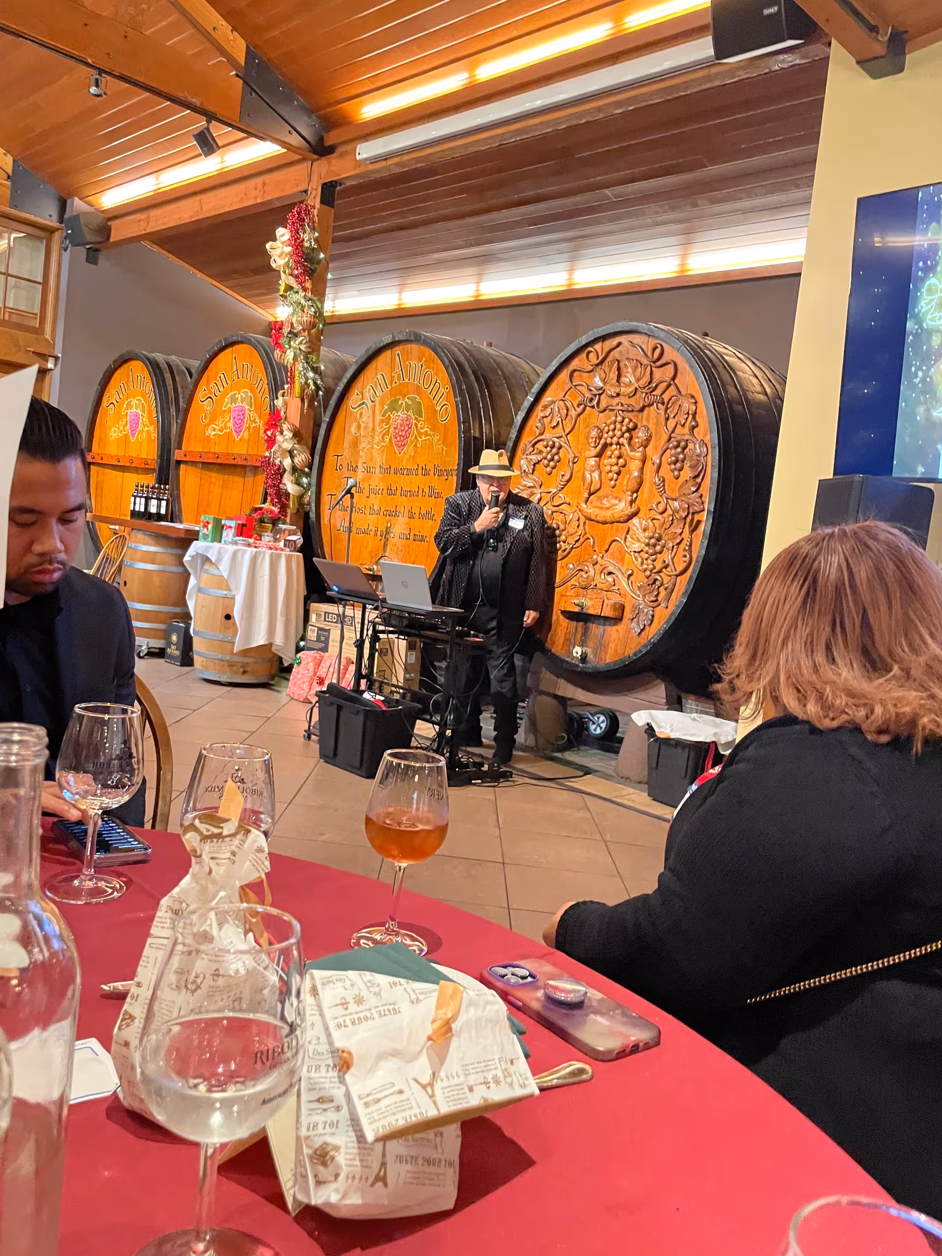 A performer wearing a hat sings into a microphone beside oversized wine barrels indoors.