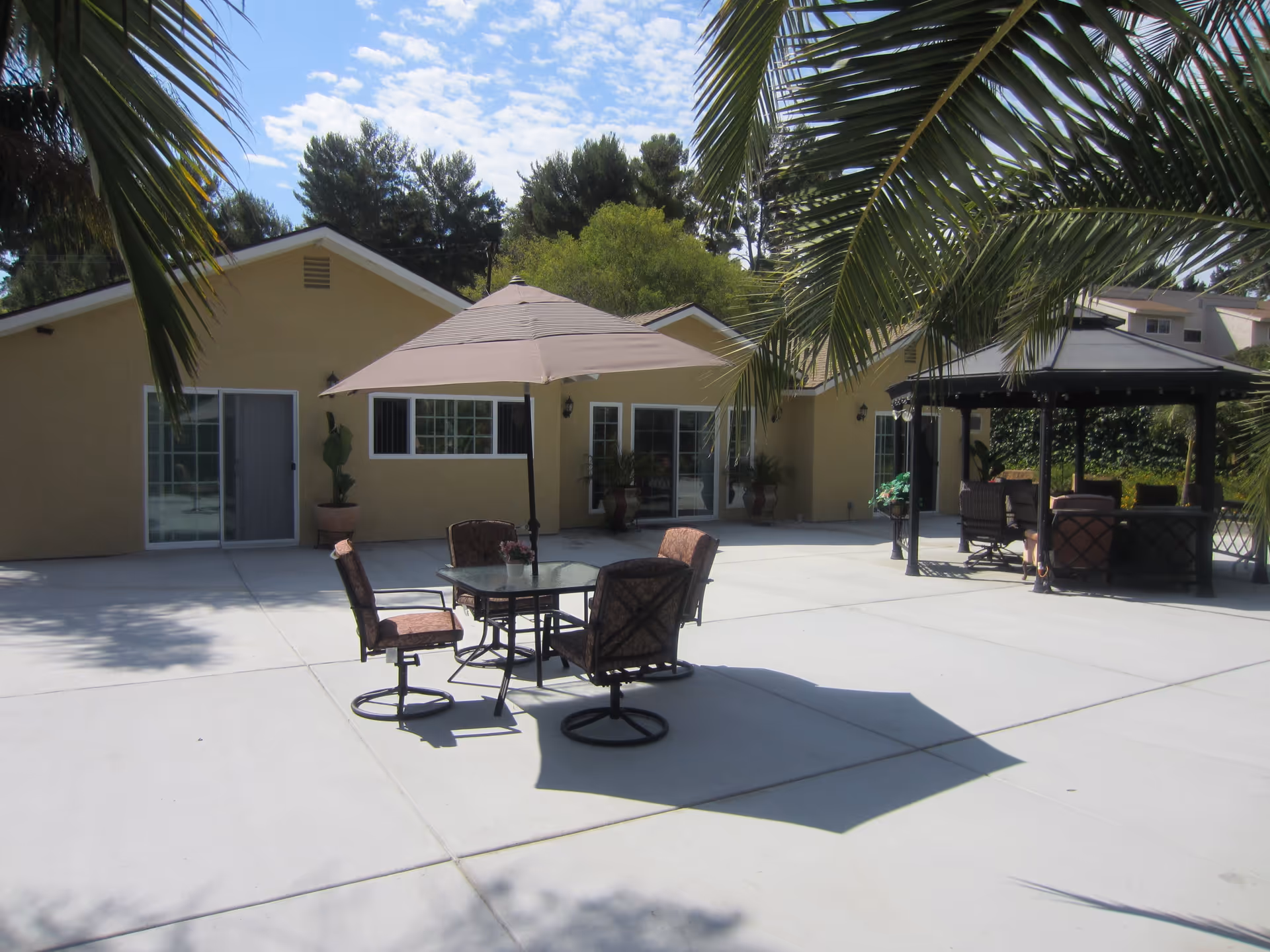 Outdoor patio area with a table and four chairs under a large beige umbrella, surrounded by palm trees. In the background, there is a yellow building with multiple windows and sliding glass doors. To the right, there is a black gazebo with additional seating underneath.