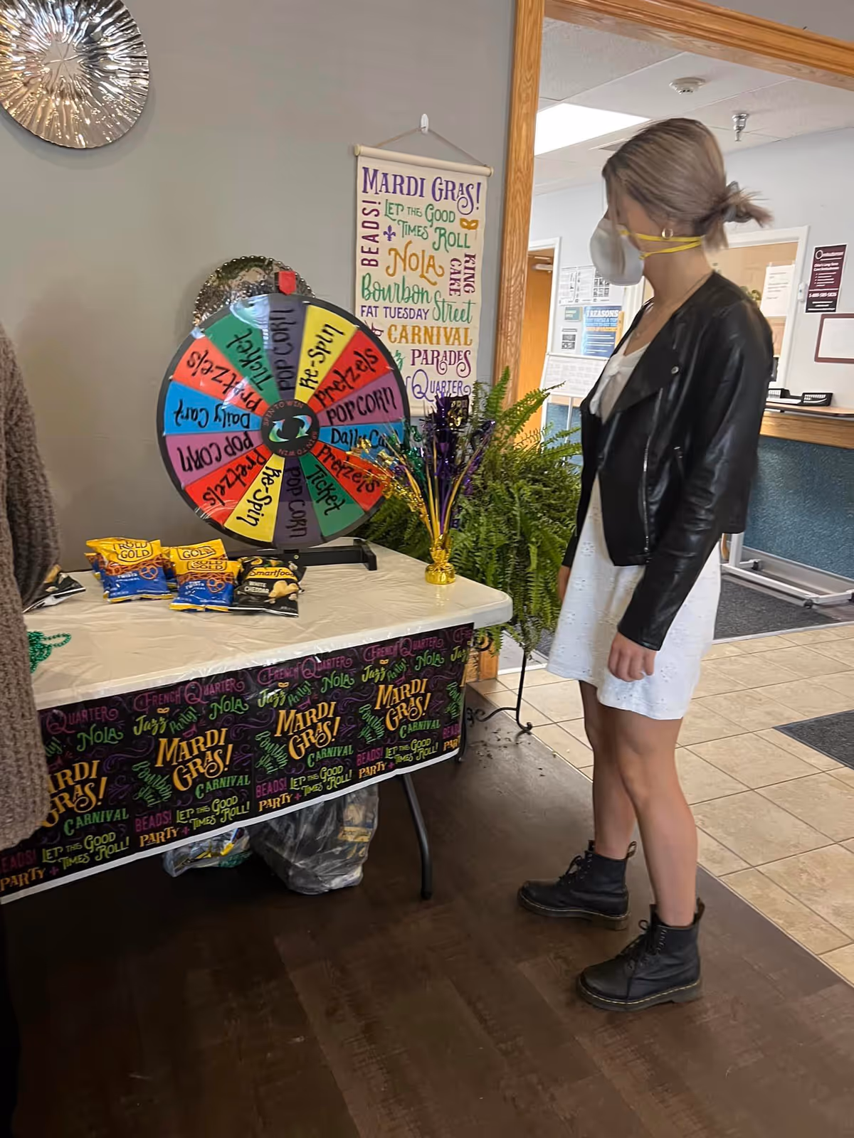 A woman wearing a white dress, black leather jacket, black boots, and a face mask stands indoors next to a table decorated with a Mardi Gras theme. On the table is a colorful prize wheel with sections labeled with prizes like popcorn, pretzels, tickets, and re-spin. The table is covered with a Mardi Gras-themed cloth and has snacks and a small decorative centerpiece. Behind the table is a wall hanging with Mardi Gras-related words and phrases.