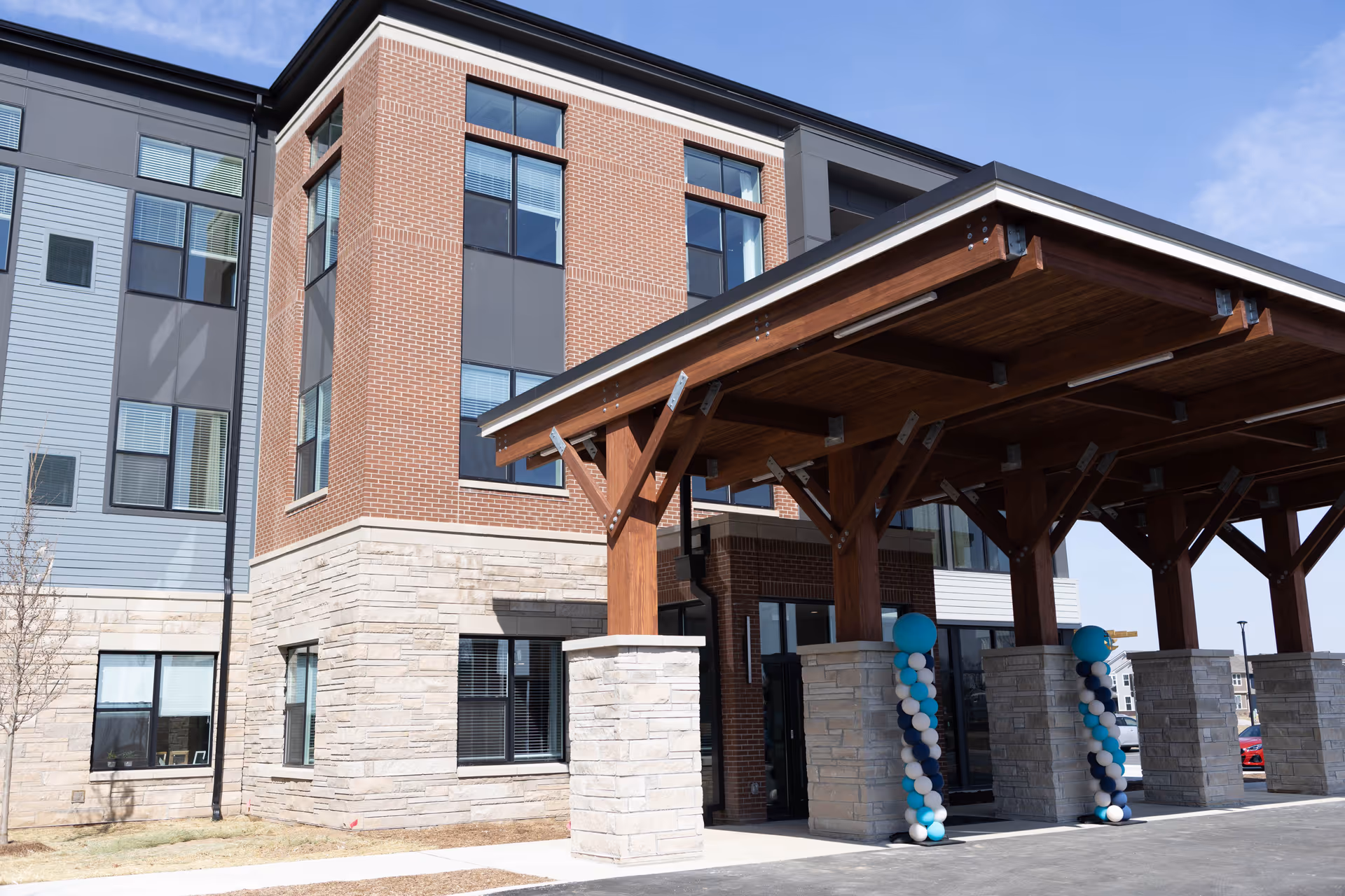 Front entrance of a multi-story brick and stone senior living building with a wooden covered driveway and decorative balloon columns.