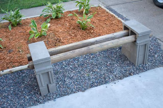 Outdoor concrete sidewalk with a wooden rail fence, mulch, and small plants.