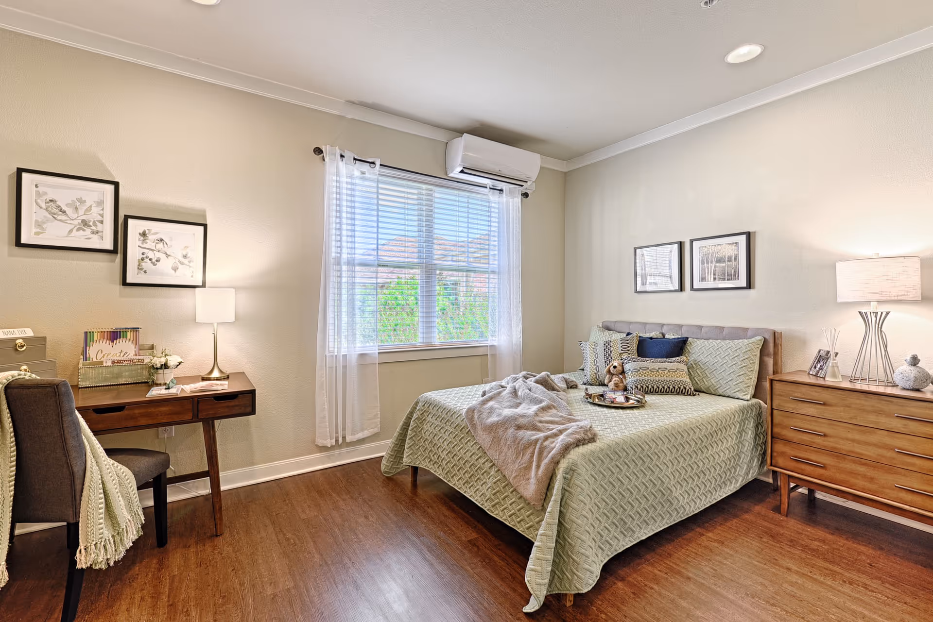 A neatly arranged senior living bedroom with bed, dresser, desk, and window.