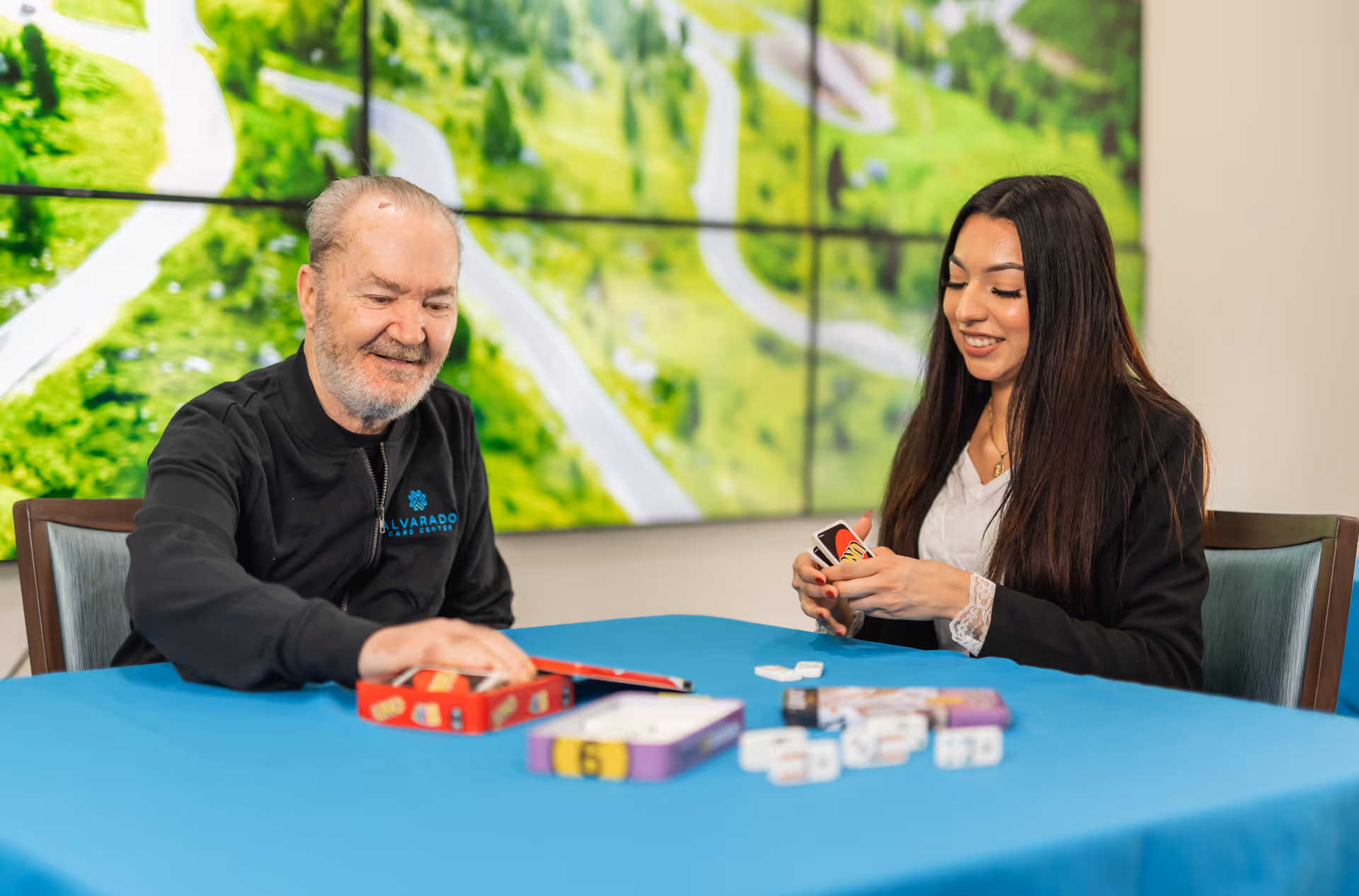 An elderly man and a young woman sitting at a table covered with a blue tablecloth, playing a card game together. The man is smiling and reaching for cards, while the woman is holding cards and smiling. Behind them is a large screen displaying an aerial view of a green landscape with roads.