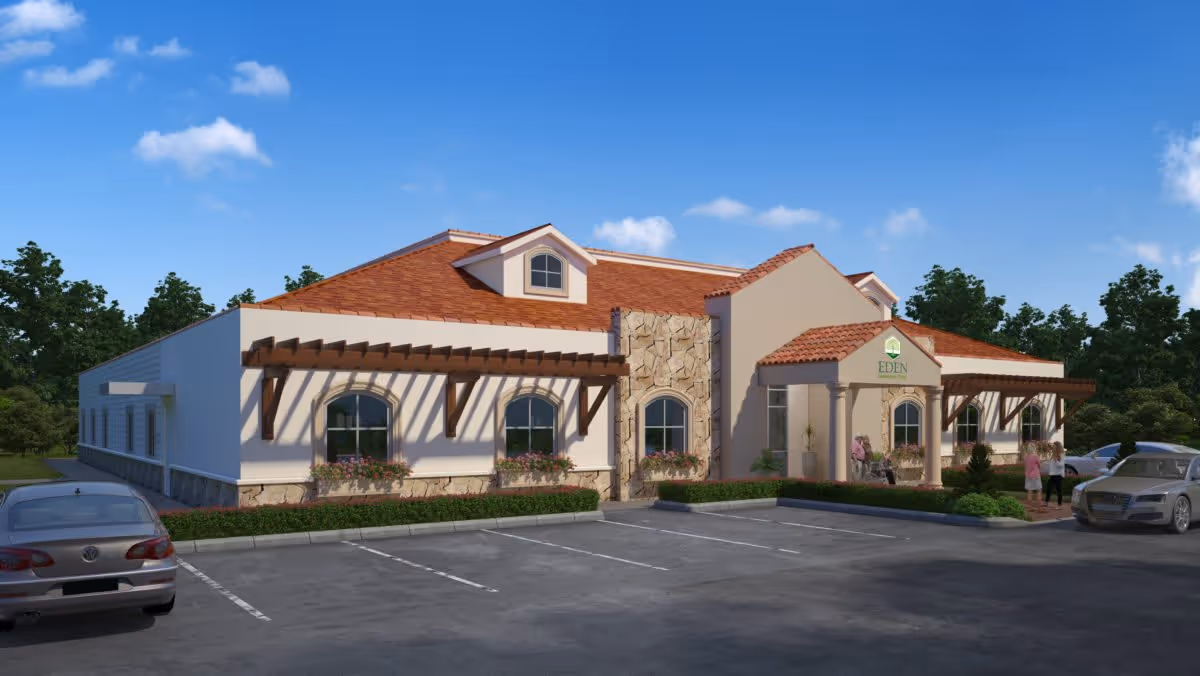Front exterior of a single-story Mediterranean-style memory care building with parked cars, landscaping, and people near the entrance.