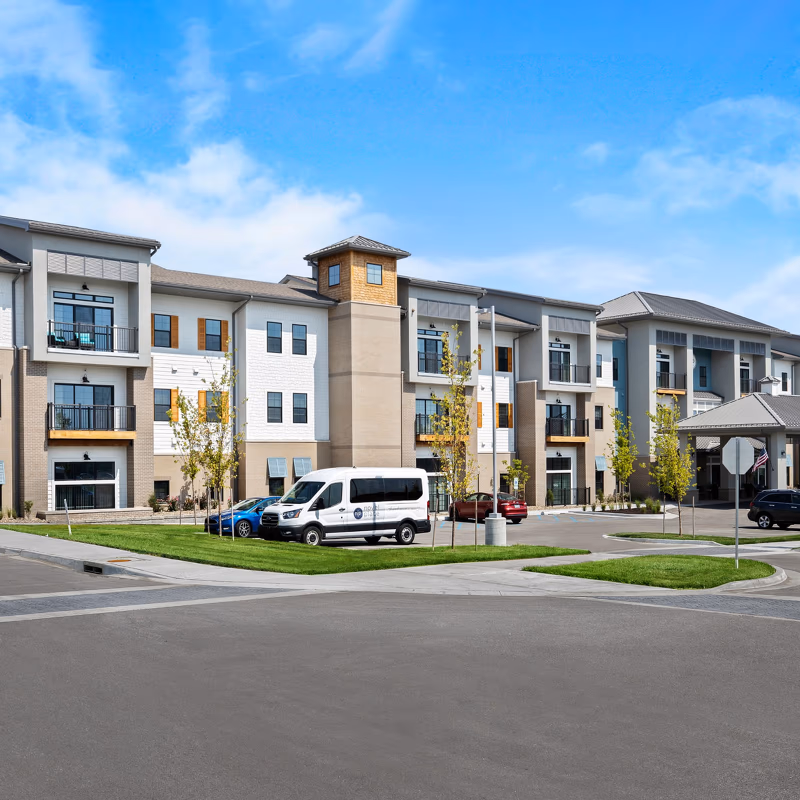 Modern three-story senior living building with balconies, an entrance canopy, parked vehicles and landscaped lawn under a blue sky.