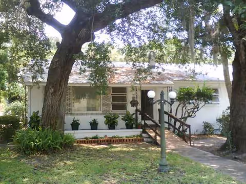 Small single-story building with ramp, stairs, potted plants, and large trees in front.