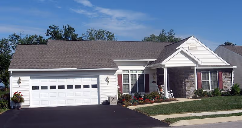 Single-story ranch-style house with an attached two-car garage, front porch with a rocking chair, and a landscaped lawn under a blue sky.