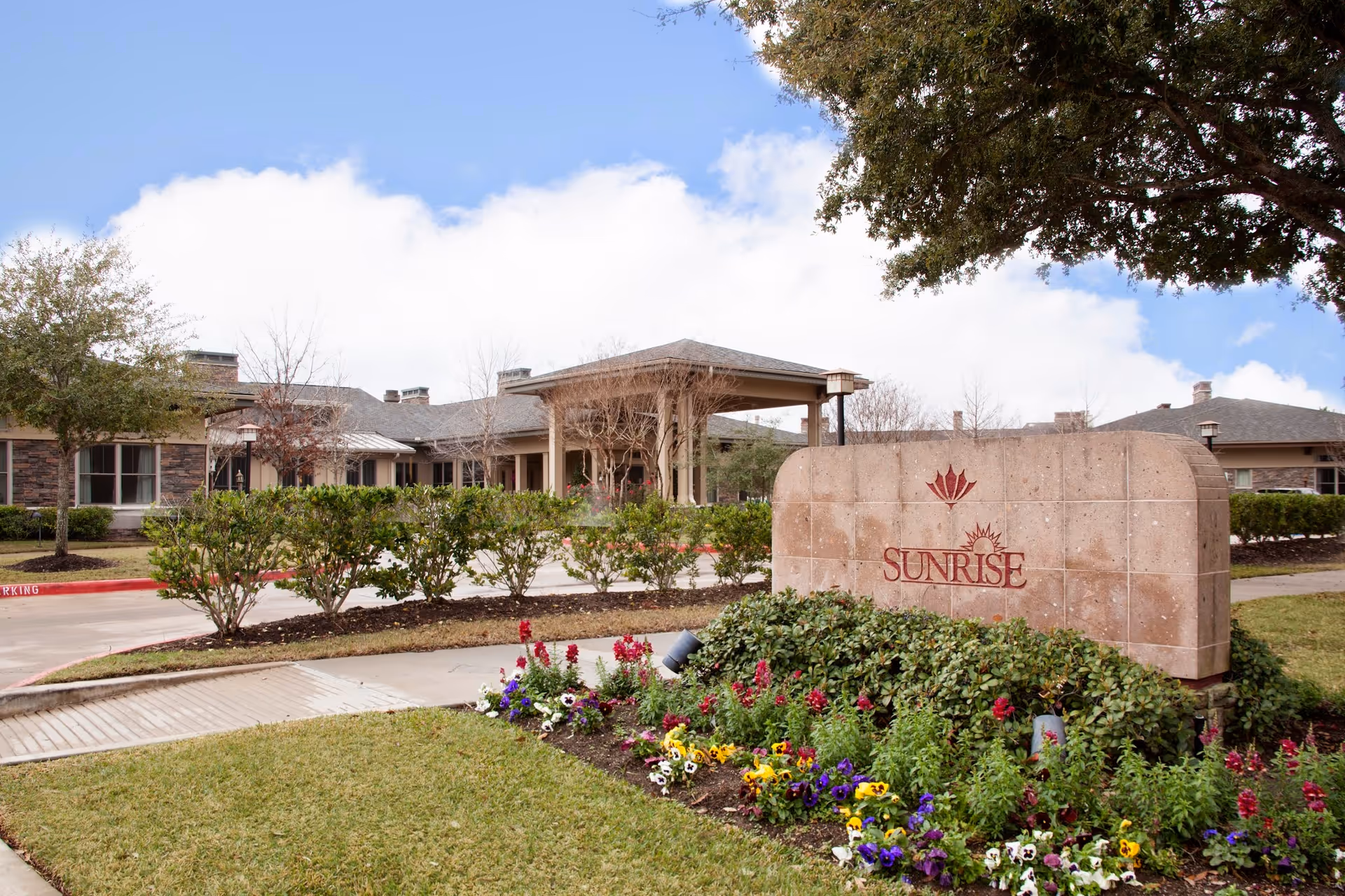 Exterior view of Sunrise of Cinco Ranch senior living facility showing a stone sign with the Sunrise logo surrounded by colorful flowers and greenery, with the building and entrance visible in the background under a partly cloudy sky.