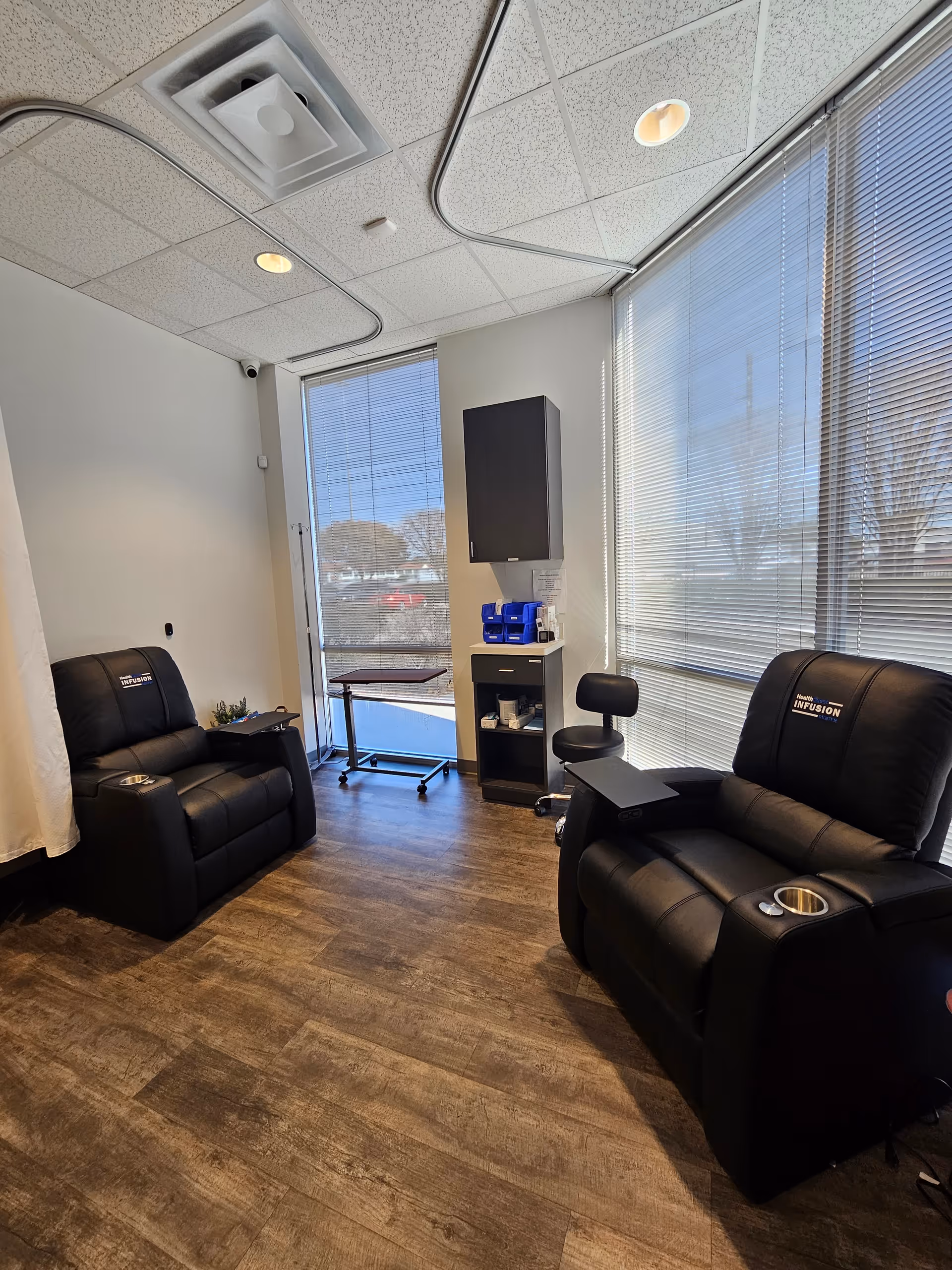 Two black recliner chairs in a bright infusion room with large windows and equipment.