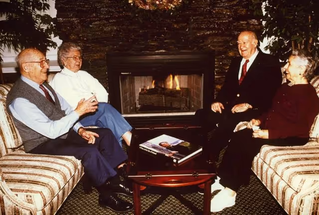Four elderly people sitting on two striped sofas facing each other in a cozy living room with a lit fireplace in the background. A wooden coffee table with books is placed between them. They appear to be engaged in conversation and smiling.