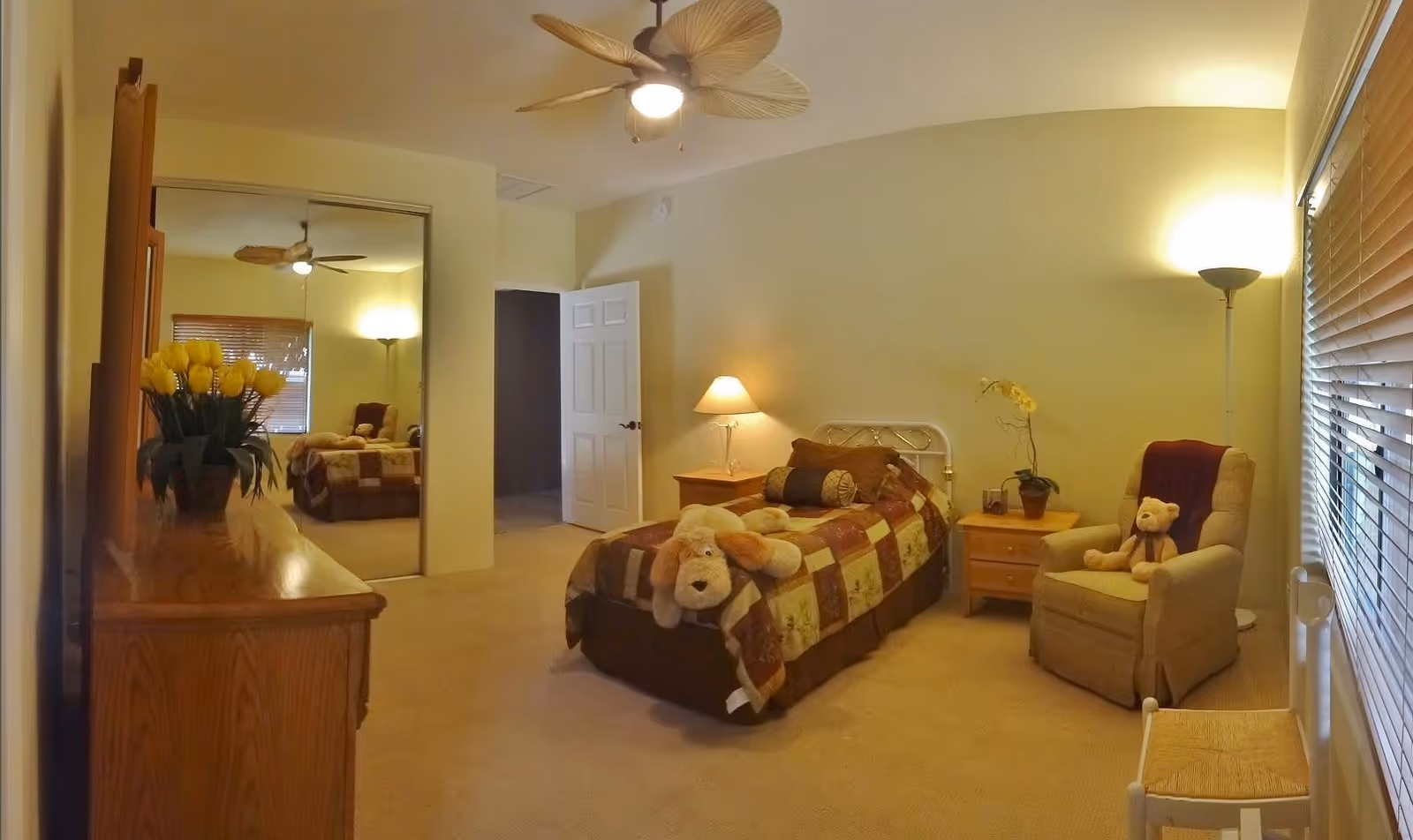 A cozy bedroom with a single bed covered in a patchwork quilt, a large stuffed dog on the bed, a wooden nightstand with a lamp, a beige armchair with a teddy bear, a floor lamp, a wooden dresser with a vase of yellow tulips, and a mirrored closet door reflecting the room.